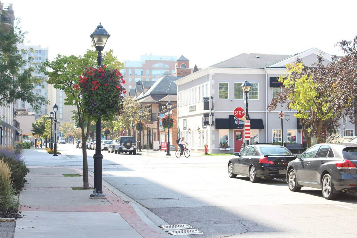 A quiet downtown street with parked cars, a cyclist, shops, trees, street lamps with hanging flower baskets, and a stop sign.