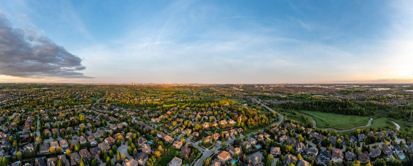 Aerial view of a suburban neighborhood with houses, trees, and winding roads during sunset, with a park and fields nearby.