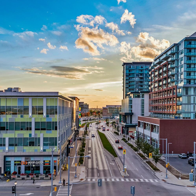City street scene during sunset with modern buildings, cars, and a partly cloudy sky.
