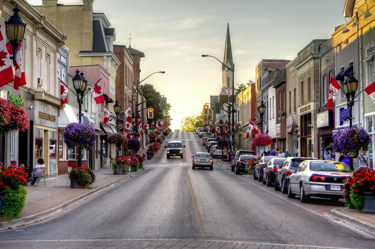 A downtown street decorated with Canadian flags and hanging flower baskets. Cars are parked along the sides of the street, with a few cars driving down the road. The buildings are colorful and lined with shops and cafes, with a church steeple visible in the background and the sun setting in the distance.