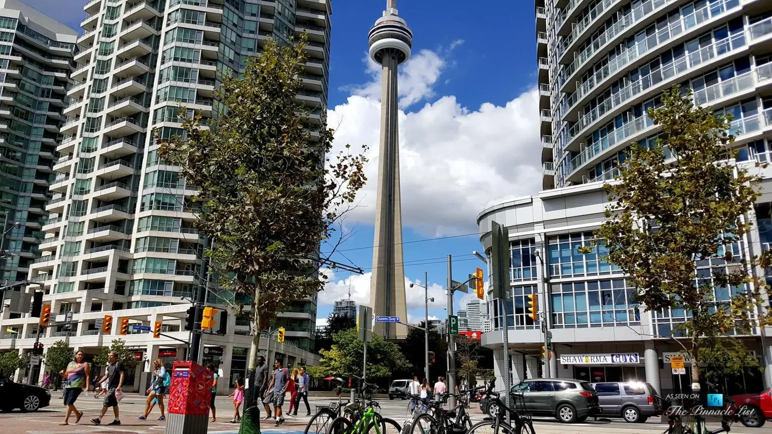 City street view featuring the CN Tower in Toronto, surrounded by modern high-rise buildings, with pedestrians, parked bicycles, and cars.