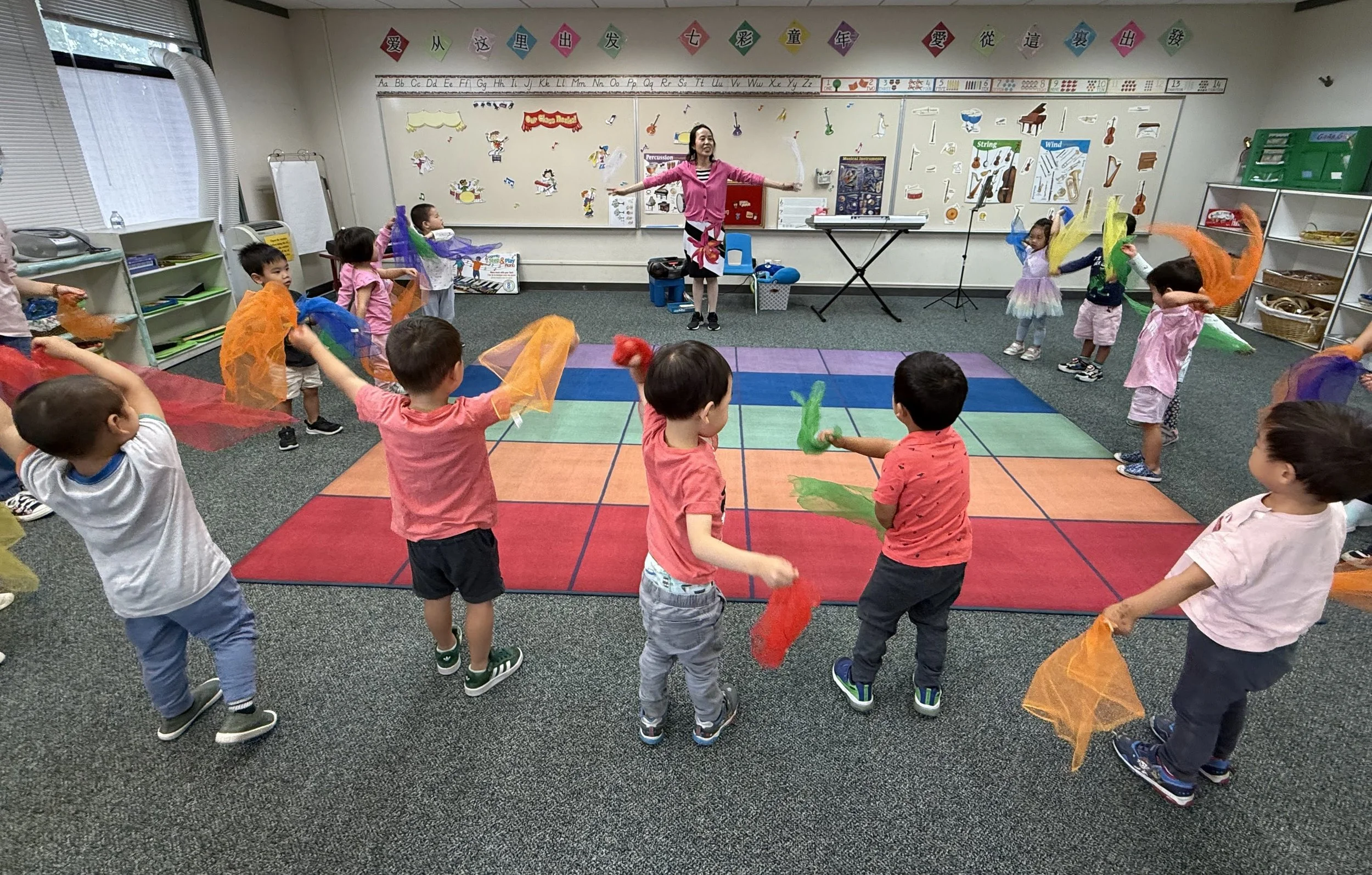 Children participating in a music or dance activity in a classroom, with a teacher leading them in a circle, holding colorful scarves or cloths.