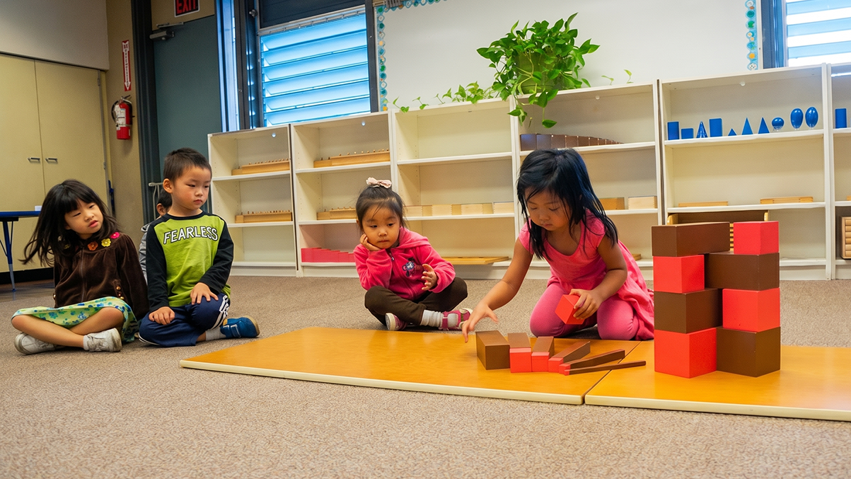 Four young children, three girls and one boy, sitting on the floor watching a girl in a pink dress stacking colorful blocks to build a tower.