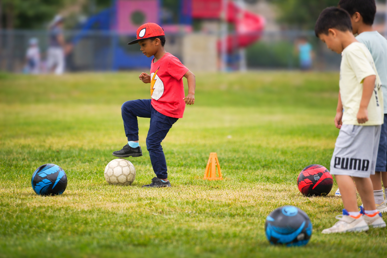 Children playing soccer on a grassy field, with soccer balls and orange cone markers, during daytime.