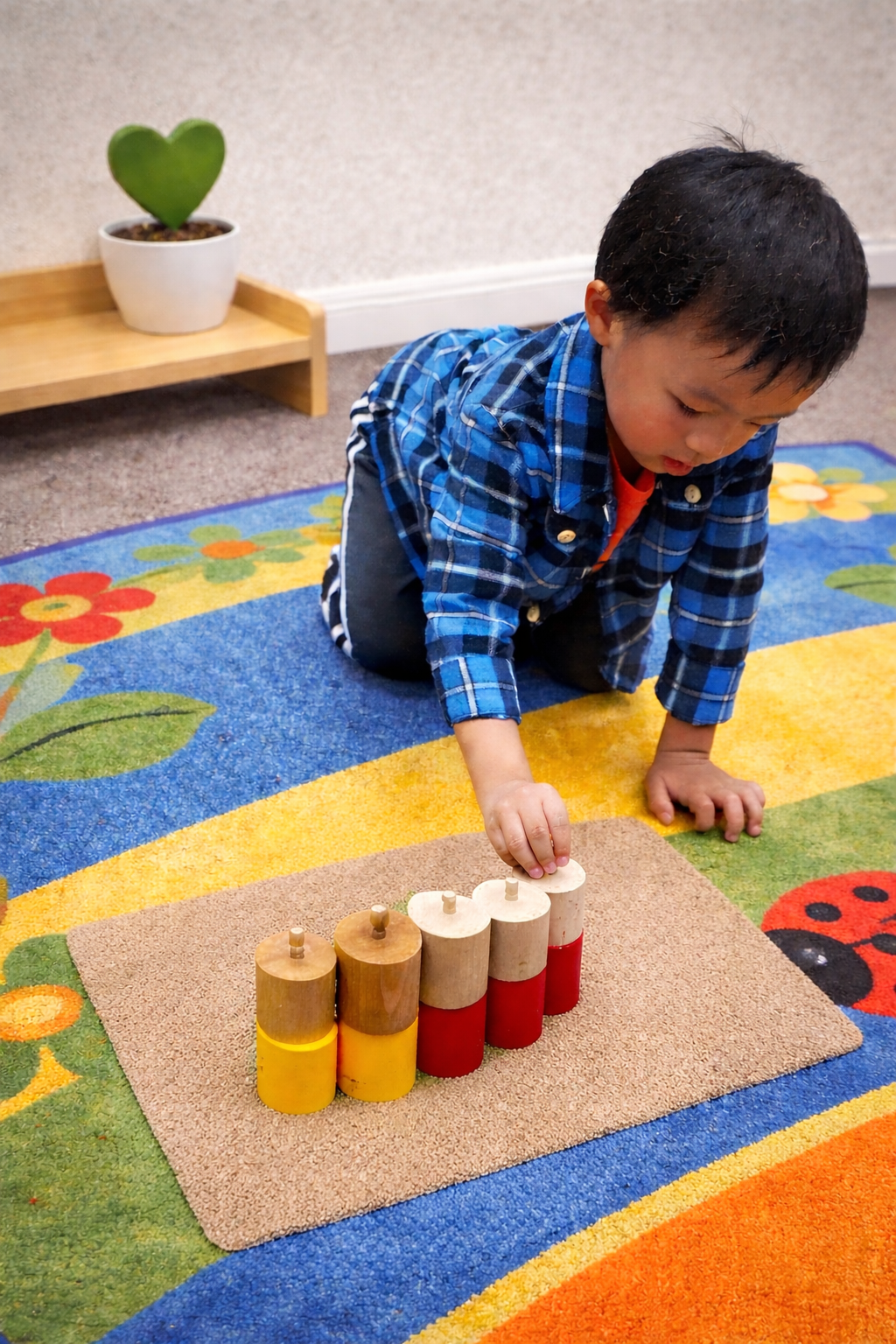 A young child in a blue plaid shirt playing on a colorful flower-themed carpet, stacking wooden cylinders.
