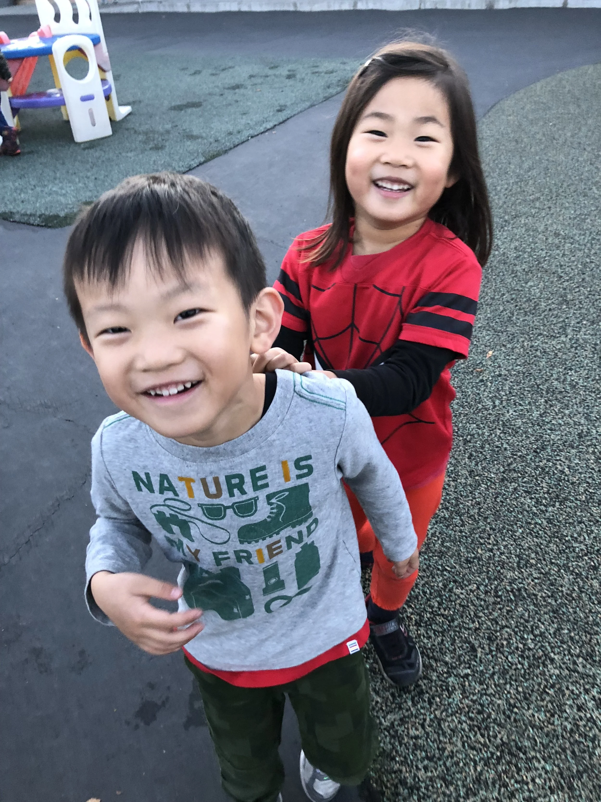 Two children, a boy and a girl, smiling and playing outdoors on a playground. The boy wears a gray shirt with green and yellow text and graphics, and the girl wears a red Spiderman-themed shirt. They appear happy and are holding hands.