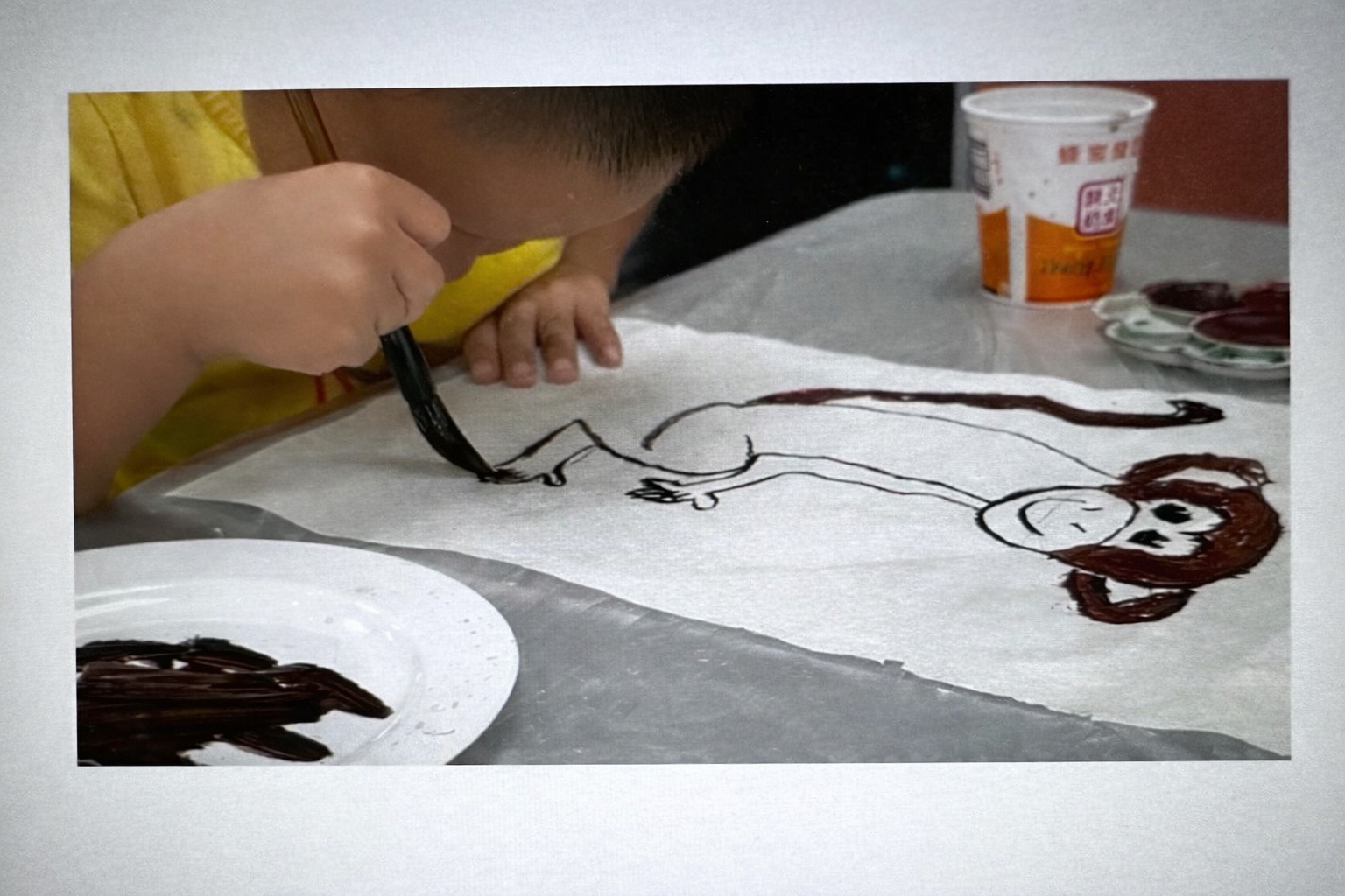 A young child uses a brush to paint a smiple monkey figure on white paper, focusing closely on the artwork at a table with paint and supplies nearby. Location is at Trust Montessori School in West San Jose. During after school program