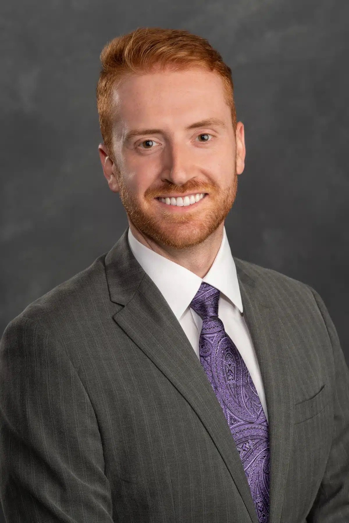 A young man with red hair and a beard wearing a dark gray suit, white shirt, and purple paisley tie, smiling against a plain dark background.
