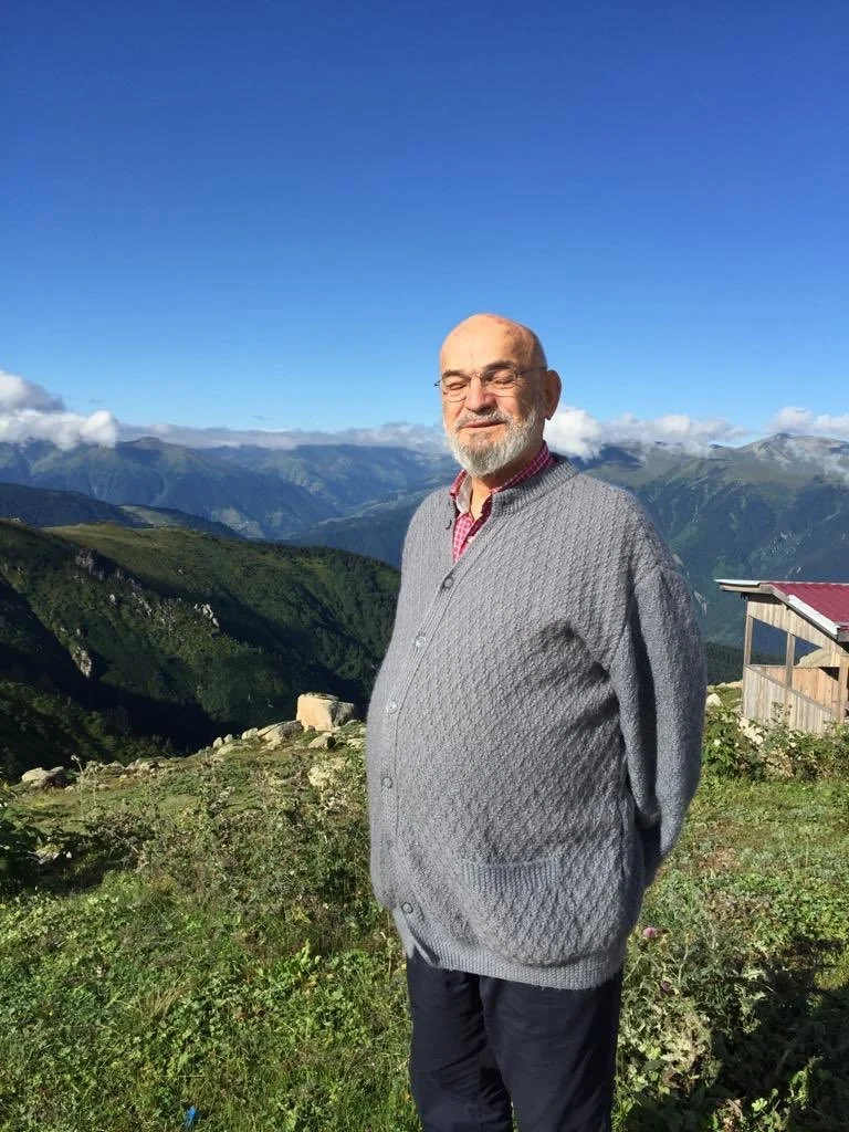 An elderly man with a grey beard and glasses standing outdoors on a grassy hillside, with mountains and a blue sky in the background.