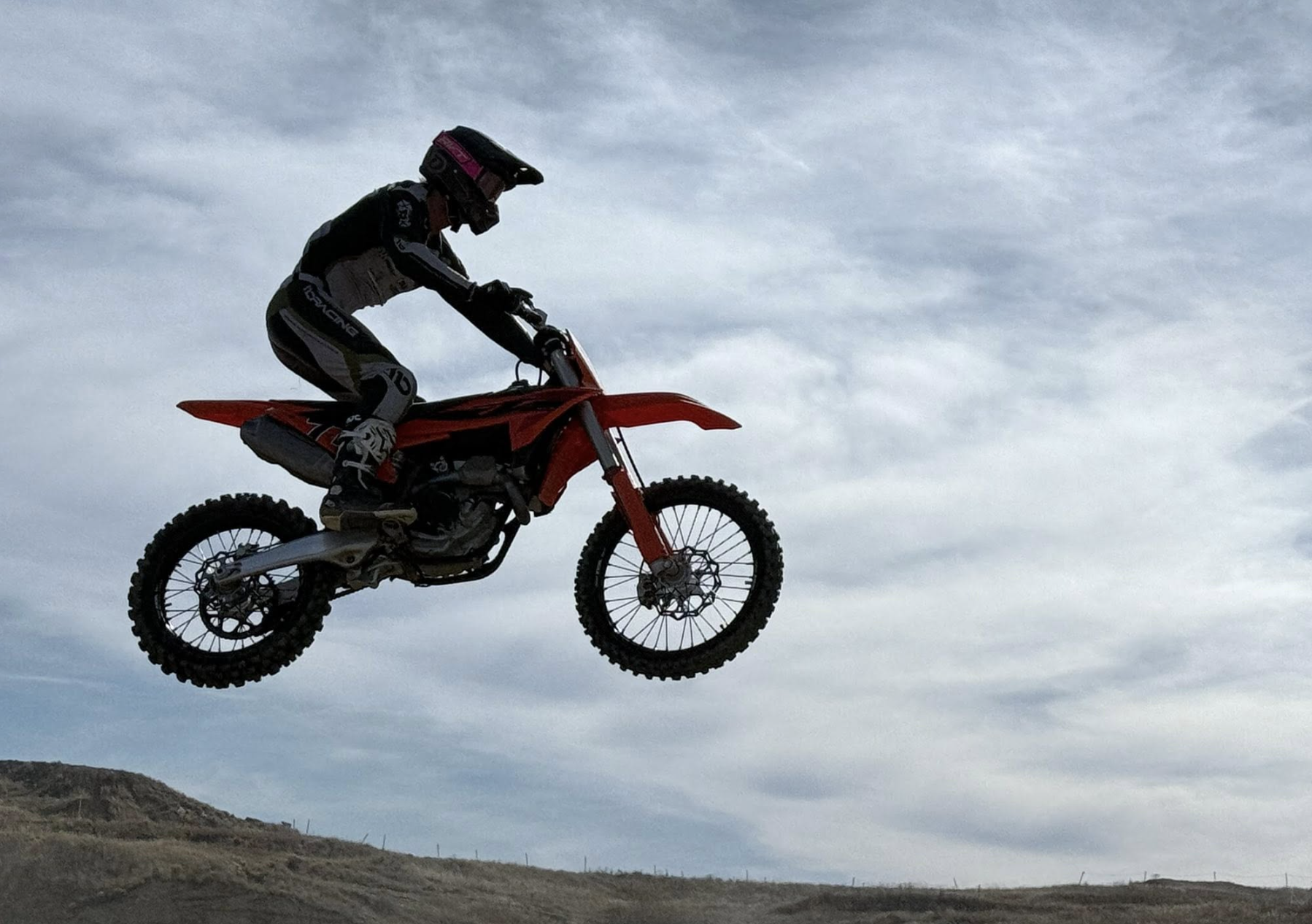A person riding a red dirt bike jumps into the air over a dirt trail with a cloudy sky in the background.