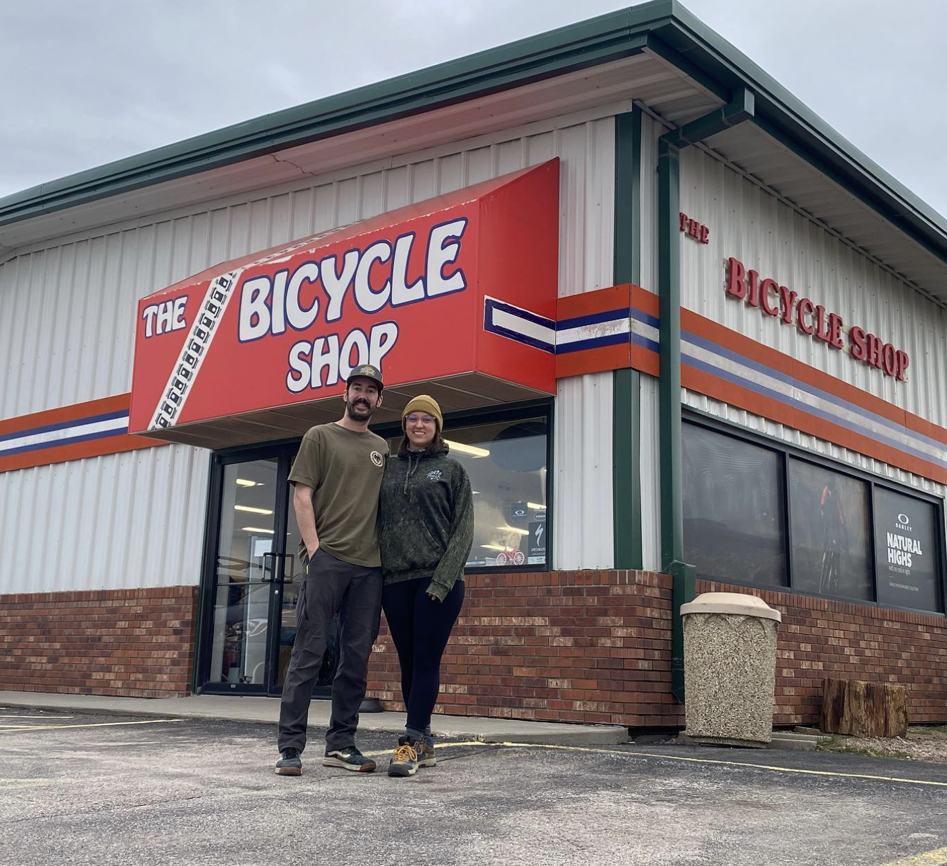 A man and woman standing outside a bicycle shop with a bright red sign, on cloudy day, in front of a brick and metal building.