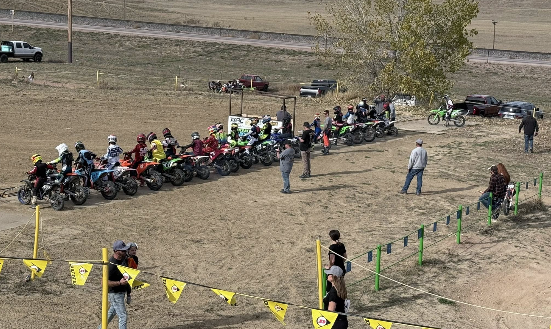 Group of motocross riders in racing gear and helmets gathered near their dirt bikes and motorcycles, with some spectators and staff around on a dirt track.