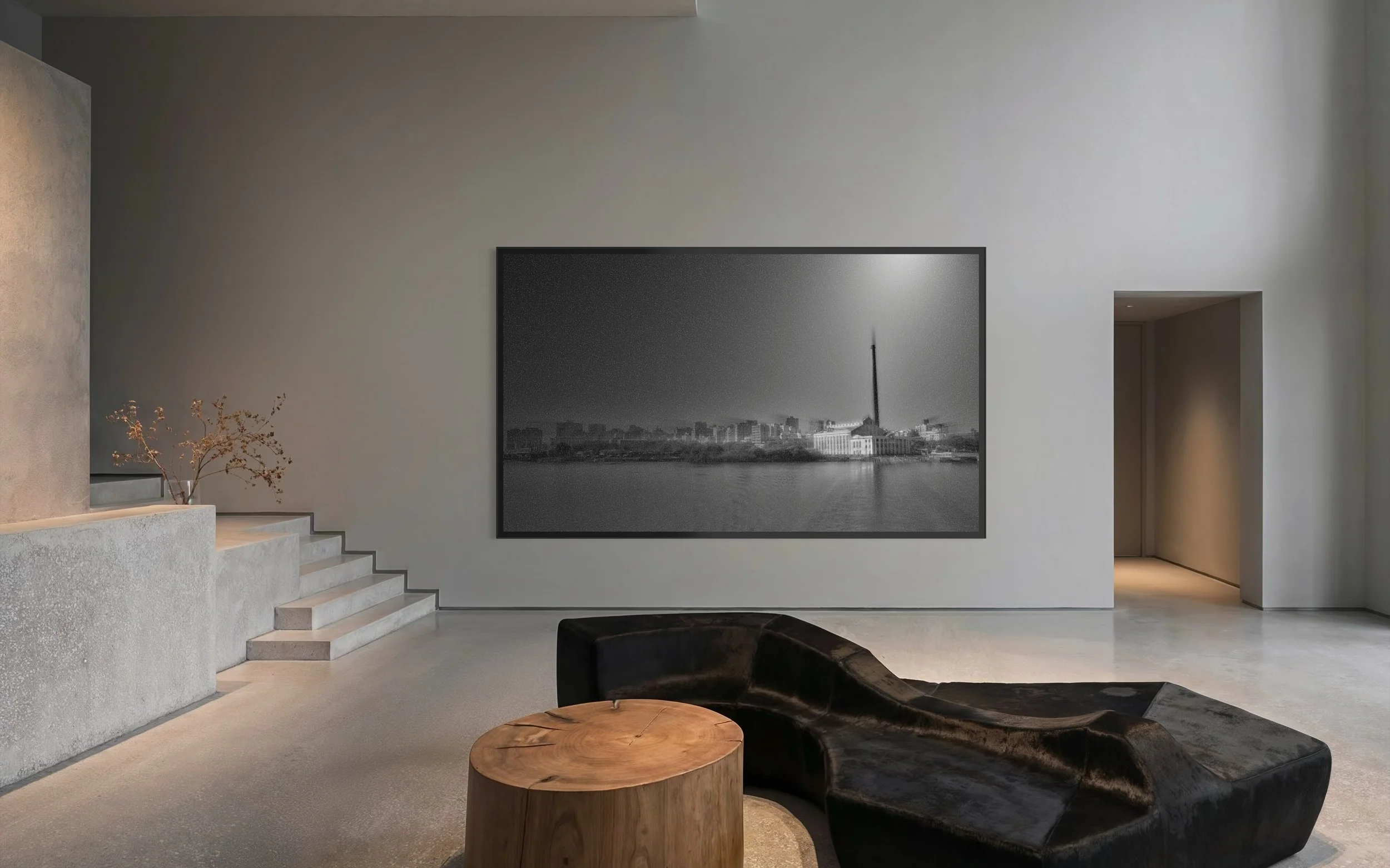 Sala de um apartamento com parede clara, obra de arte preta e branca de um rio com prédios ao fundo, um sofá preto irregular, uma mesa redonda de madeira e uma escada de concreto ao lado