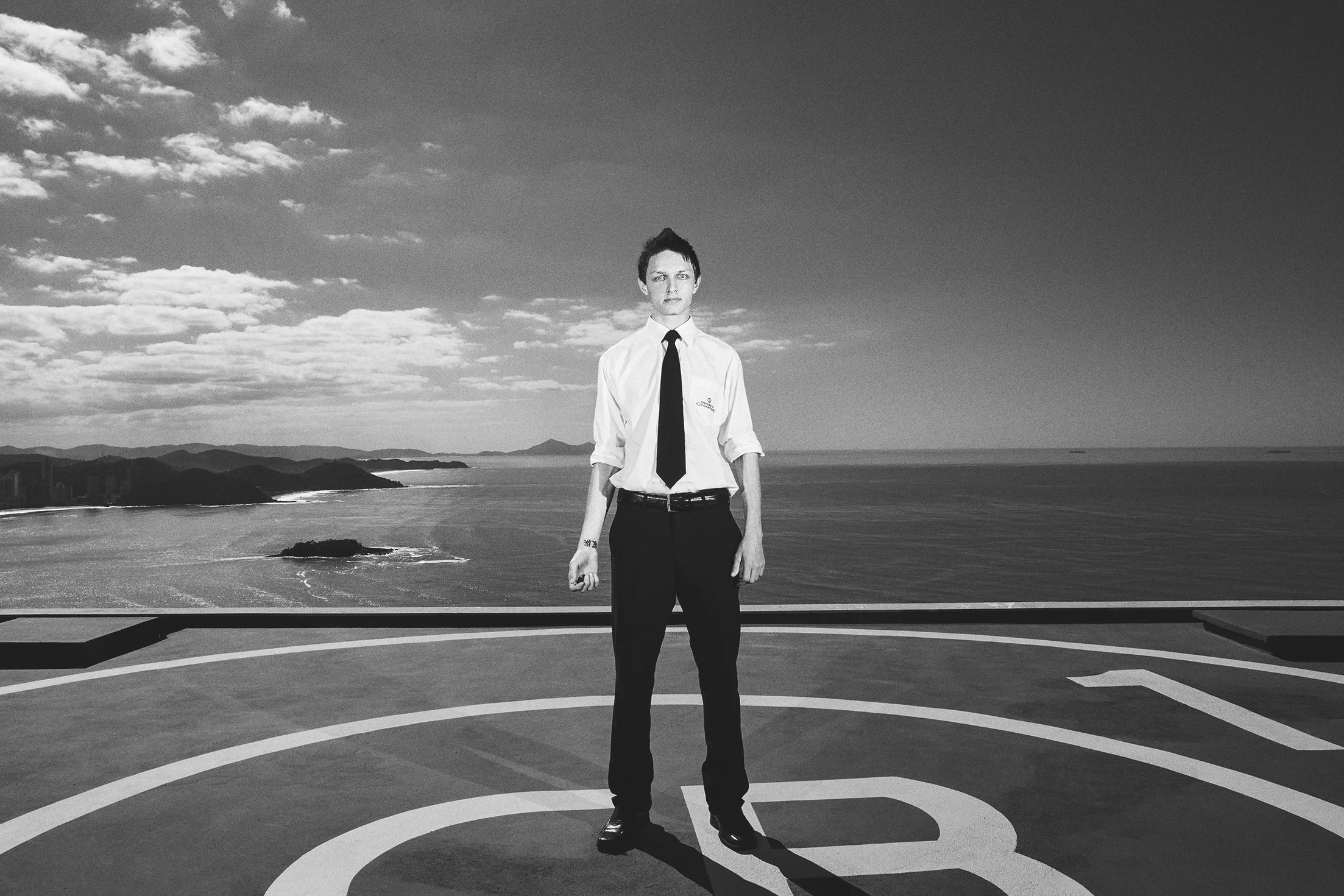 Pessoa jovem usando roupa formal, com camisa branca e gravata preta, em um heliponto ao ar livre com vista para o mar e o horizonte