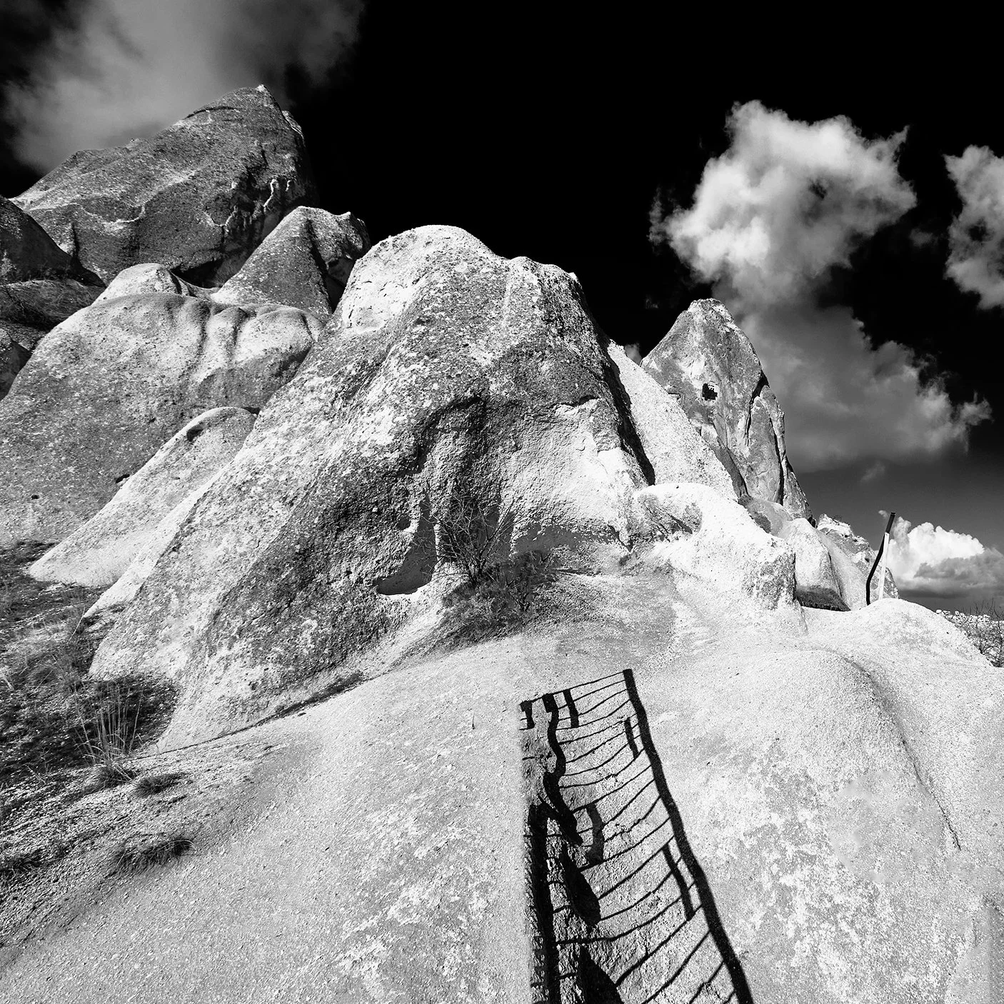 Vista de uma formação rochosa sob céu nublado em preto e branco, com uma escada de ferro suspensa na frente.