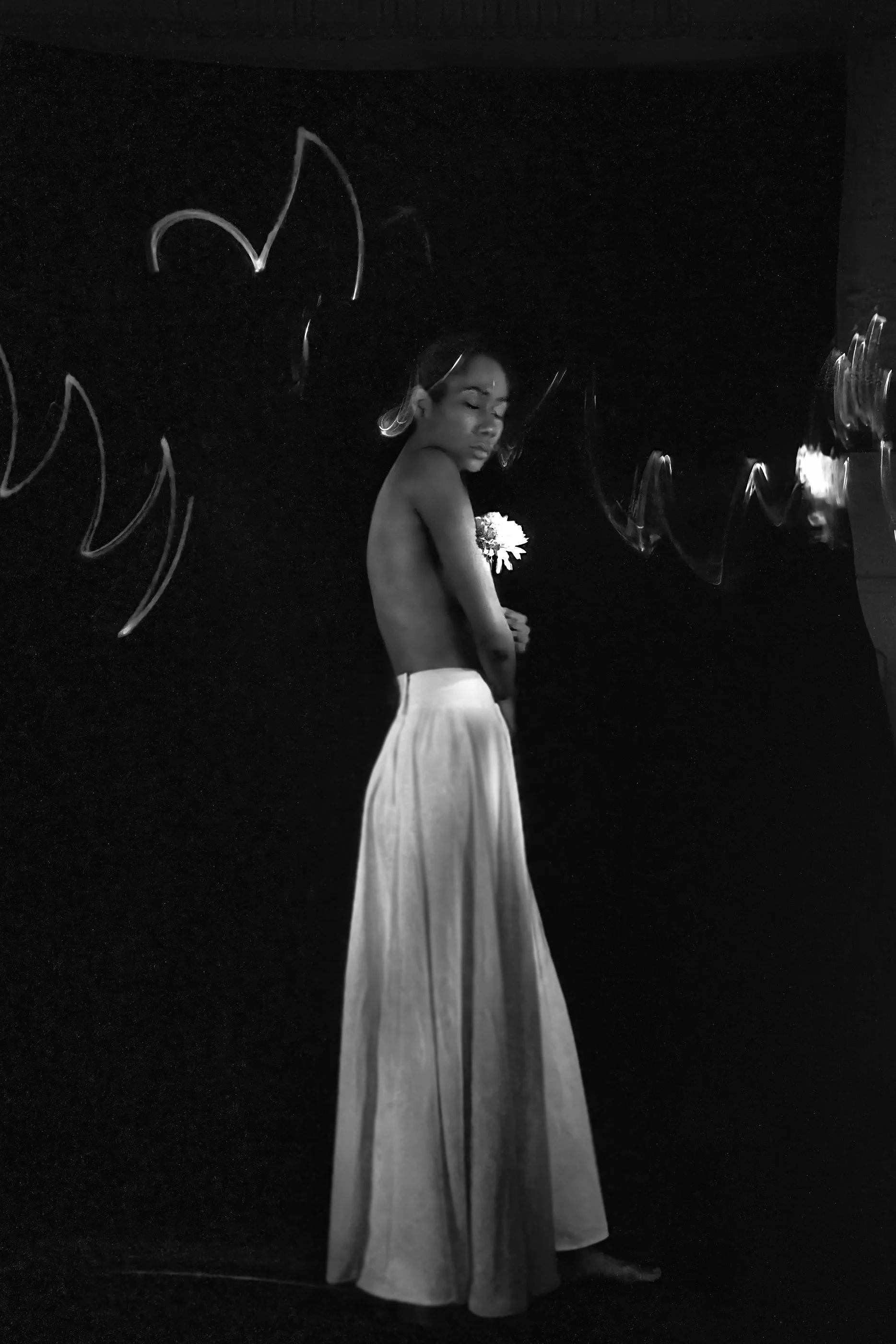 Imagem em preto e branco de uma mulher nua com saia comprida, segurando uma flor, com uma parede escura ao fundo.