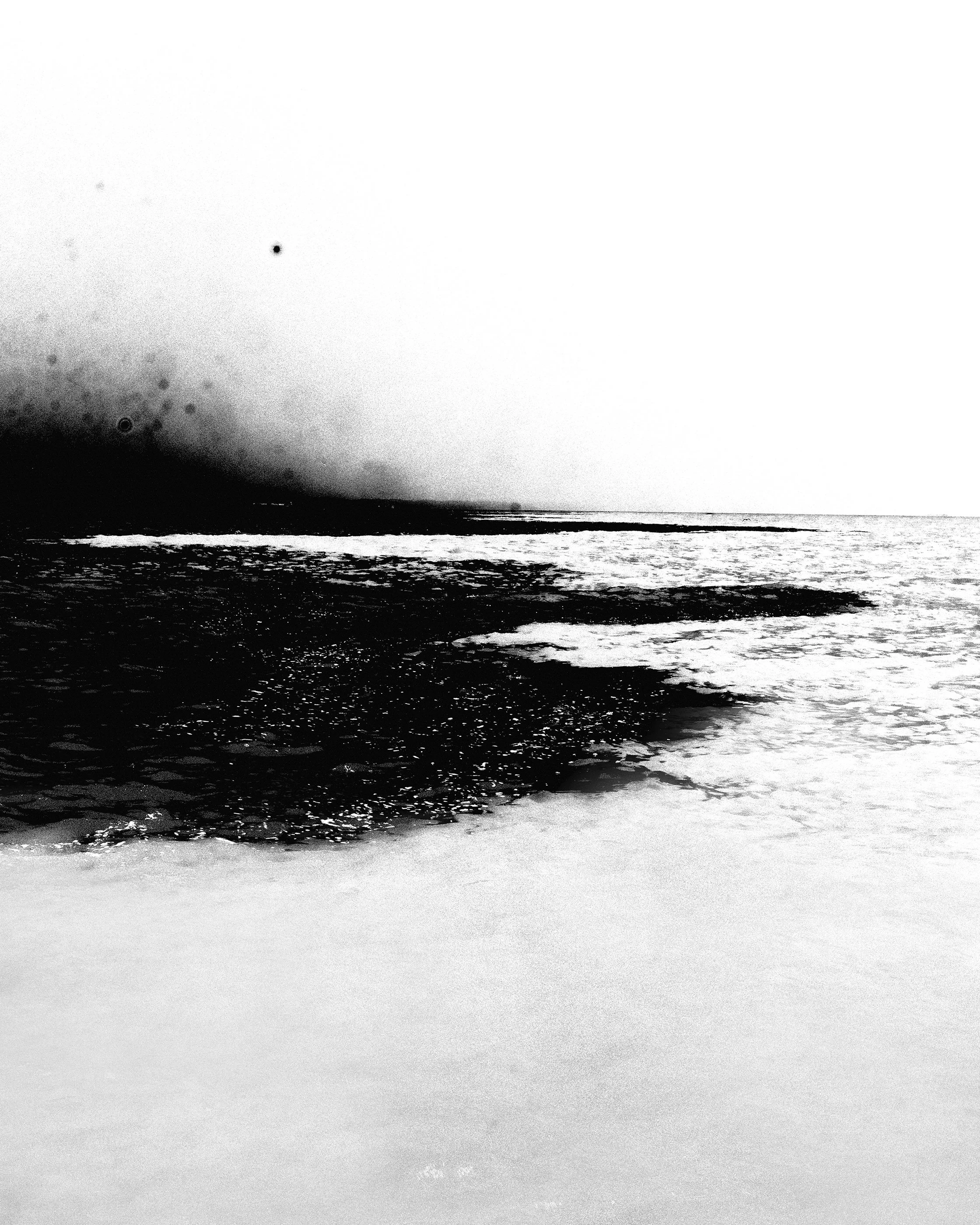 Praia deserta com ondas e céu claro, em preto e branco.
