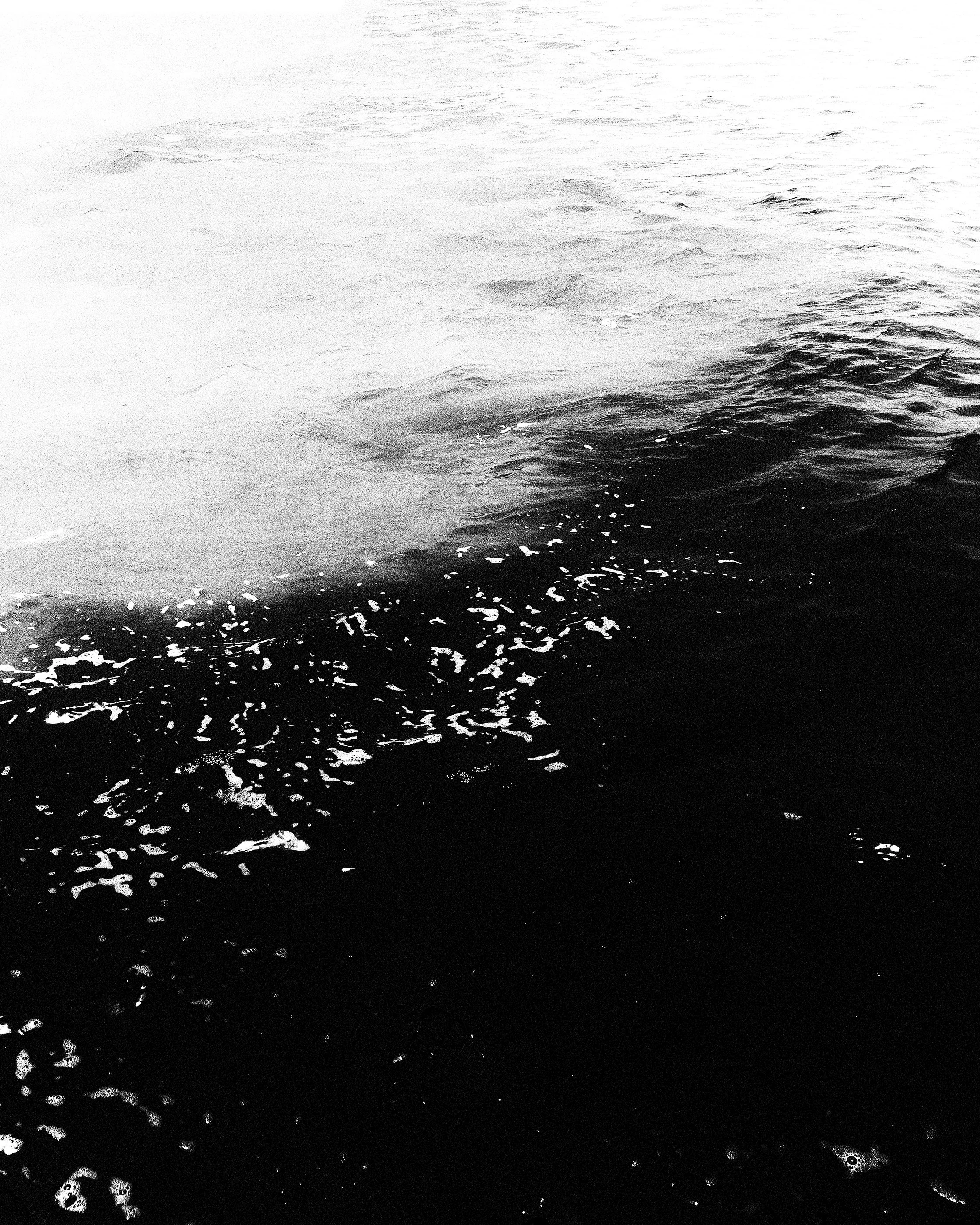Imagem em preto e branco de uma porção de água, com destaque para as áreas de água mais clara à esquerda e as áreas mais escuras à direita, com ondas e espuma na superfície.