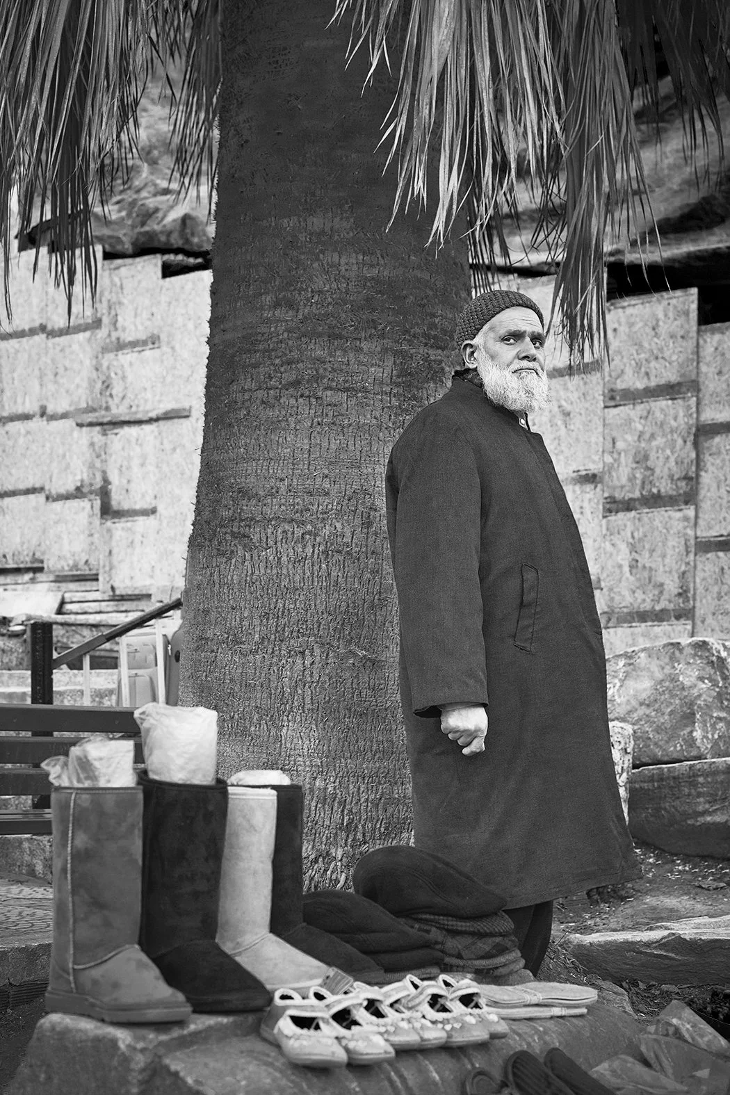 Homem idoso com barba branca, vestindo um casaco escuro, ao lado de uma árvore, com sapatos e roupas em uma mesa à sua frente, em um ambiente ao ar livre.