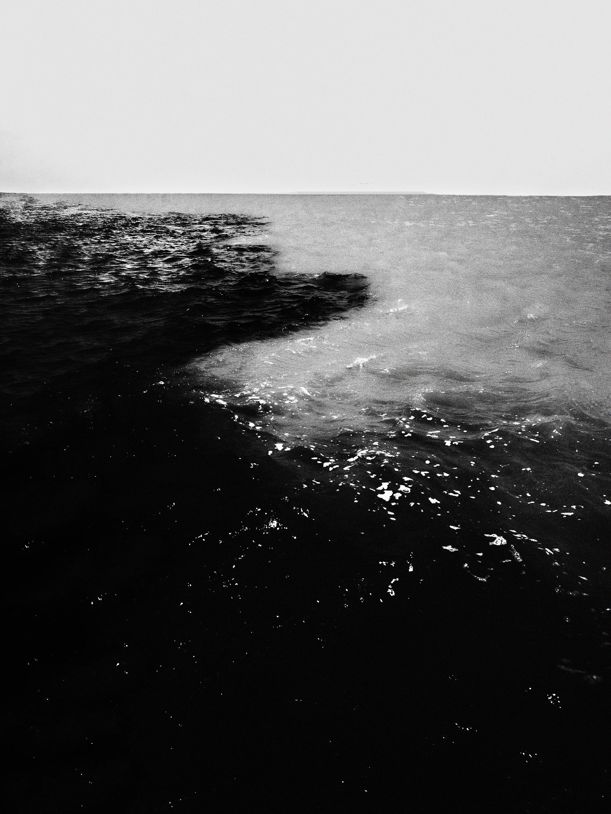 Foto em preto e branco de uma linha de separação entre a água clara do mar e a água escura, possivelmente o rastro de uma embarcação, com o horizonte ao fundo.
