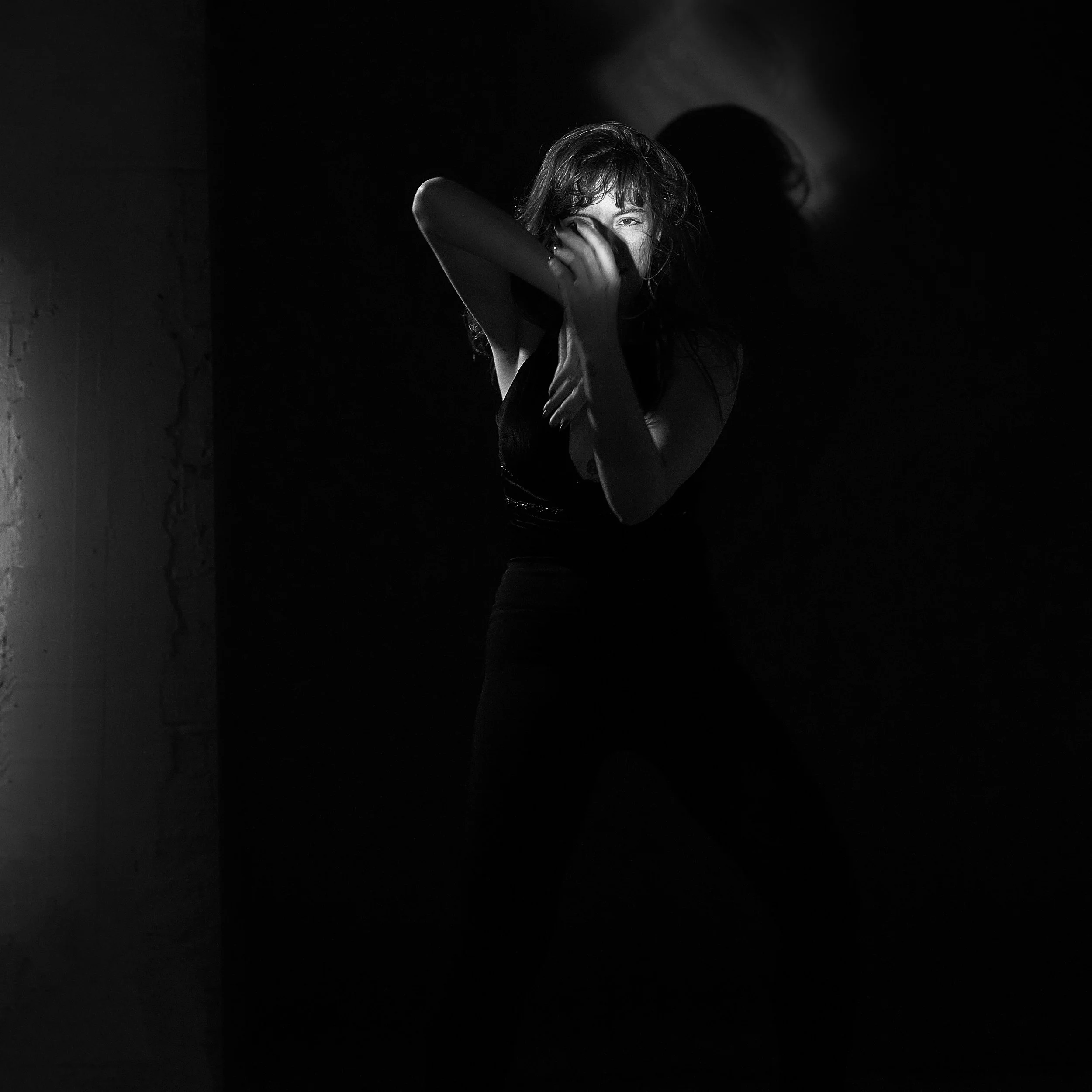 Mulher posando em uma parede, com sombra e iluminação dramática, usando roupas pretas.