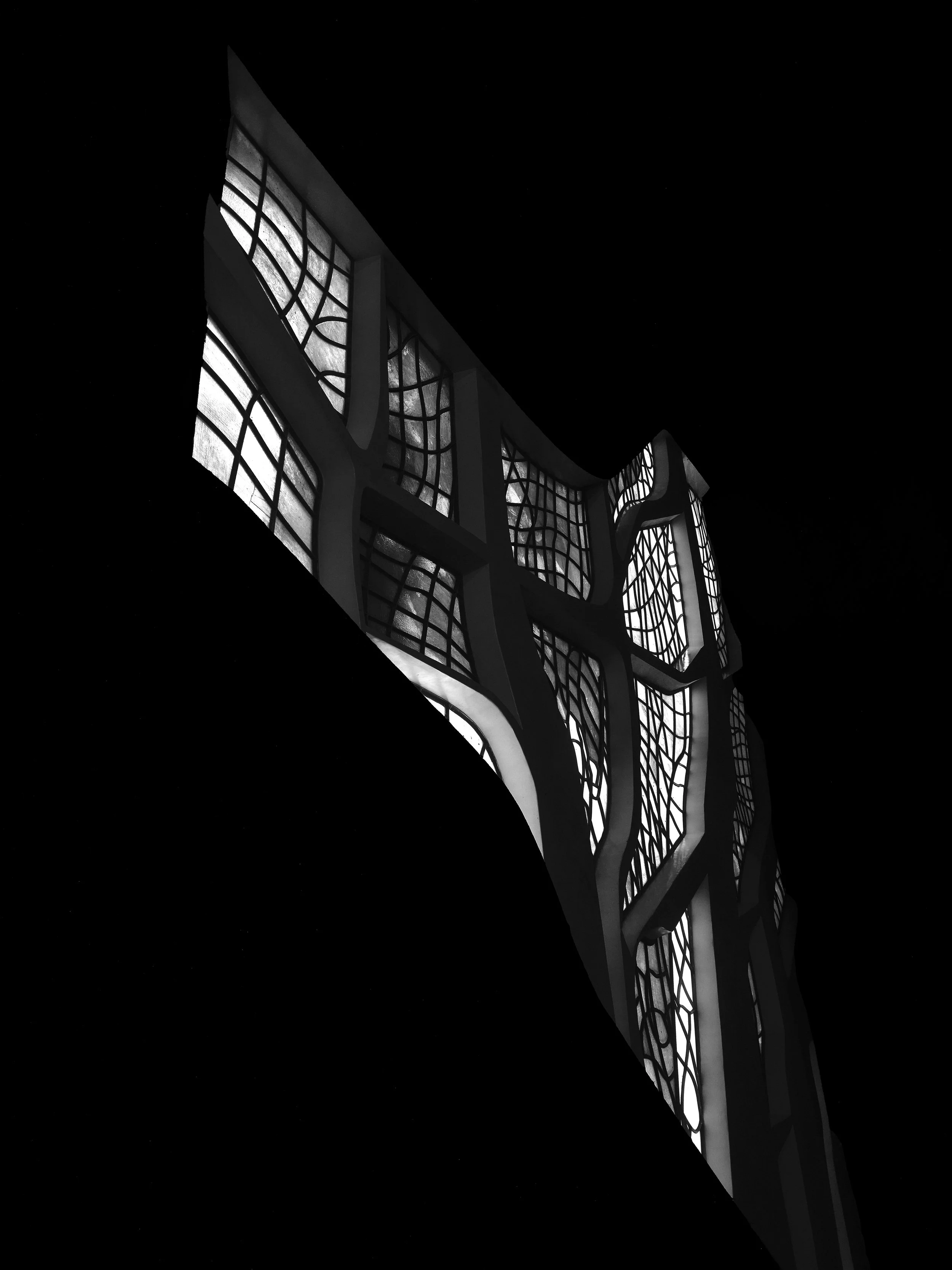 Detalhe de uma estrutura arquitetônica com janelas de vidro com padrões geométricos, vista de baixo para cima em contraste de preto e branco.