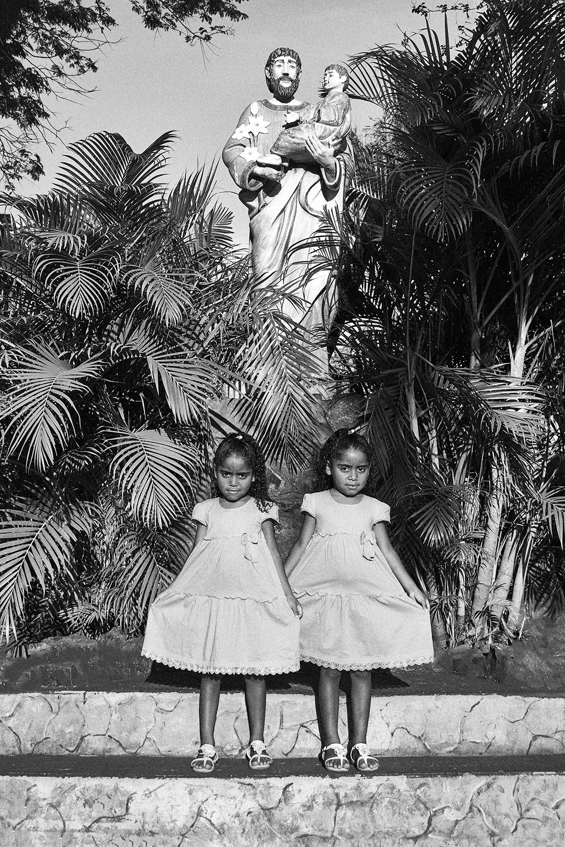 Duas meninas de vestido claro posando na frente de uma escultura religiosa de Jesus segurando uma criança, com arbustos ao fundo.