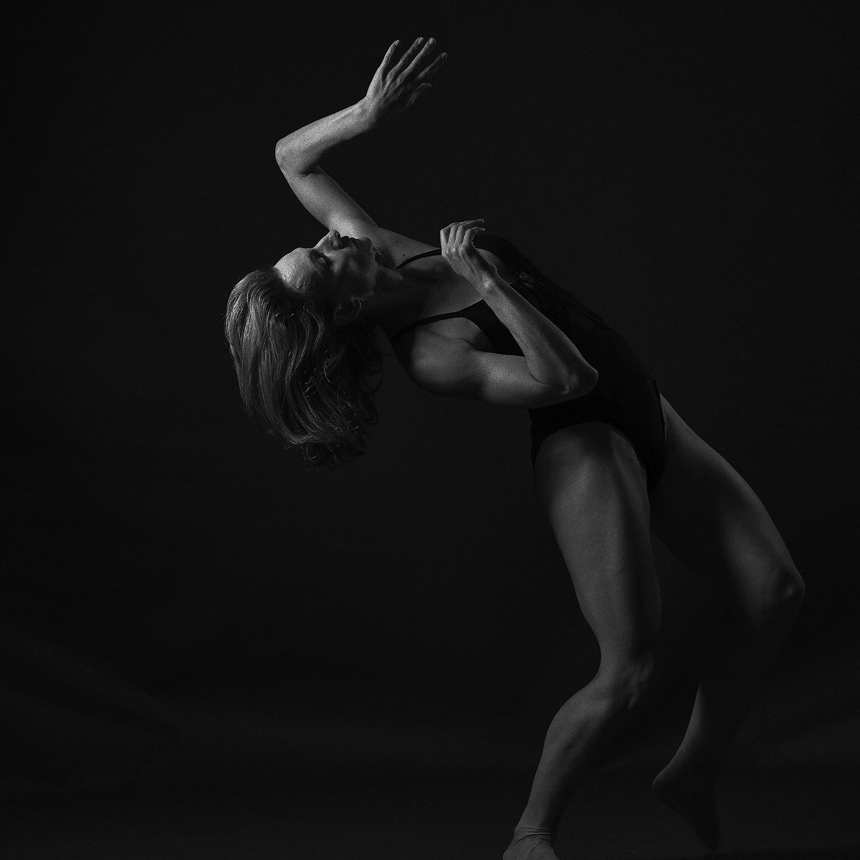 Imagem em preto e branco de uma mulher posando artisticamente com uma expressão séria, com cabelo solto, vestindo uma roupa escura, contra um fundo escuro.