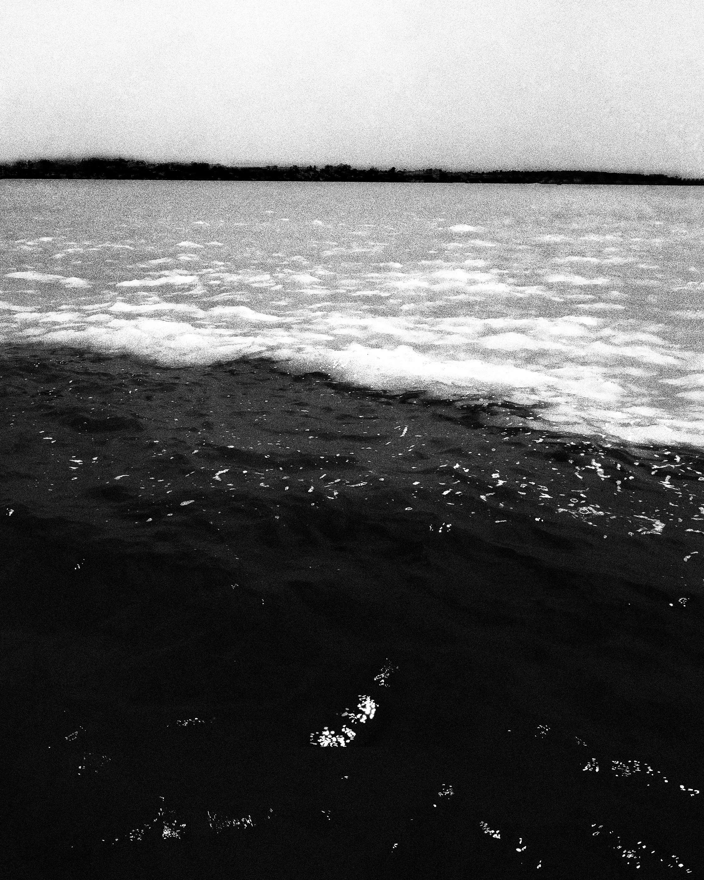 Foto em preto e branco de um corpo d'água, com ondas suaves na superfície e uma linha de terra ou árvores ao fundo.