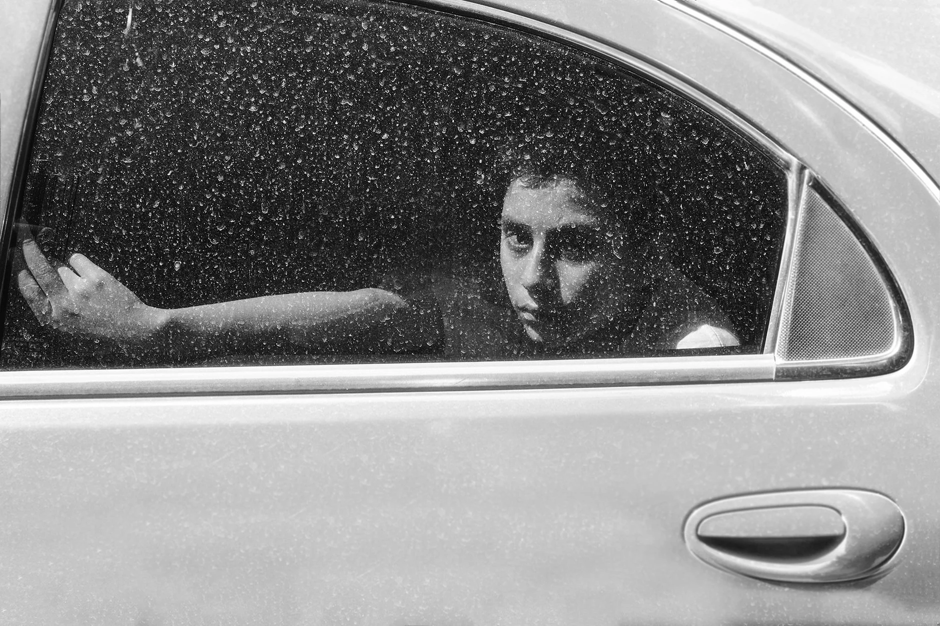 Mulher com expressão pensativa, observando através de janela de carro suja de gotas de água.