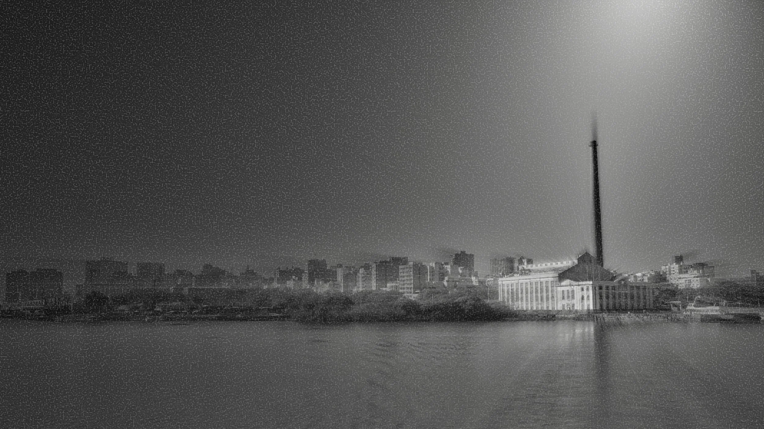 Imagem em preto e branco da cidade com um rio na frente, prédios ao fundo e uma construção com uma torre alta na direita.