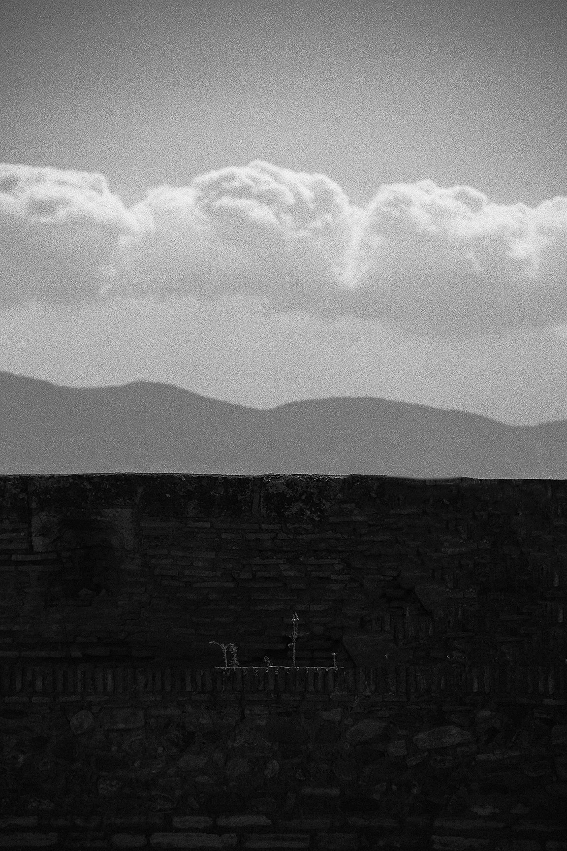 Paisagem em tons de cinza com nuvens no céu, montanhas ao fundo e uma parede de pedra na parte inferior da imagem.