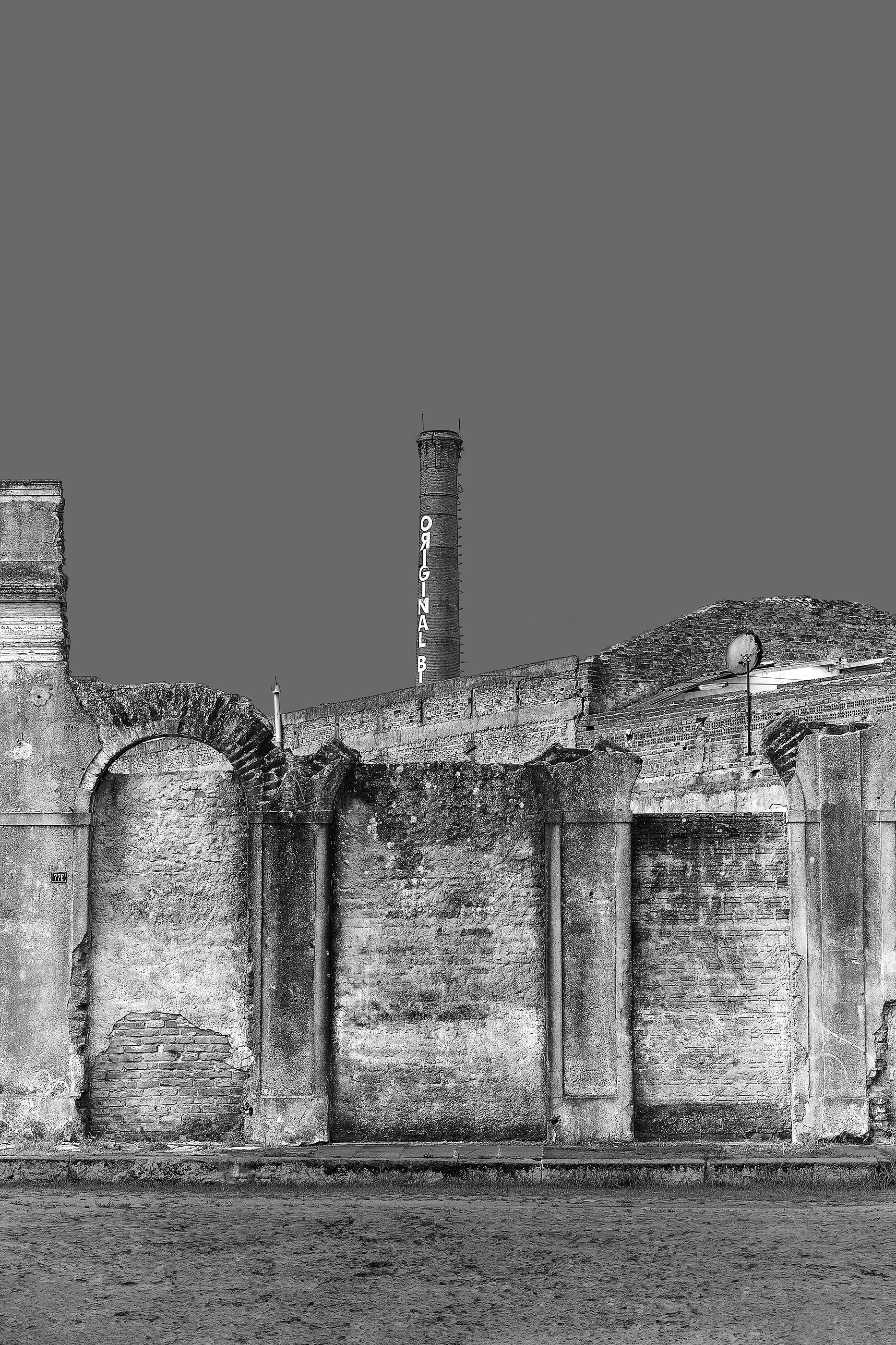 Imagem em preto e branco de uma parede antiga de alvenaria com um arco, com uma chaminé ao fundo com a palavra 'ORIGINAL' escrita nela, e um satélite na parte superior direita da parede.