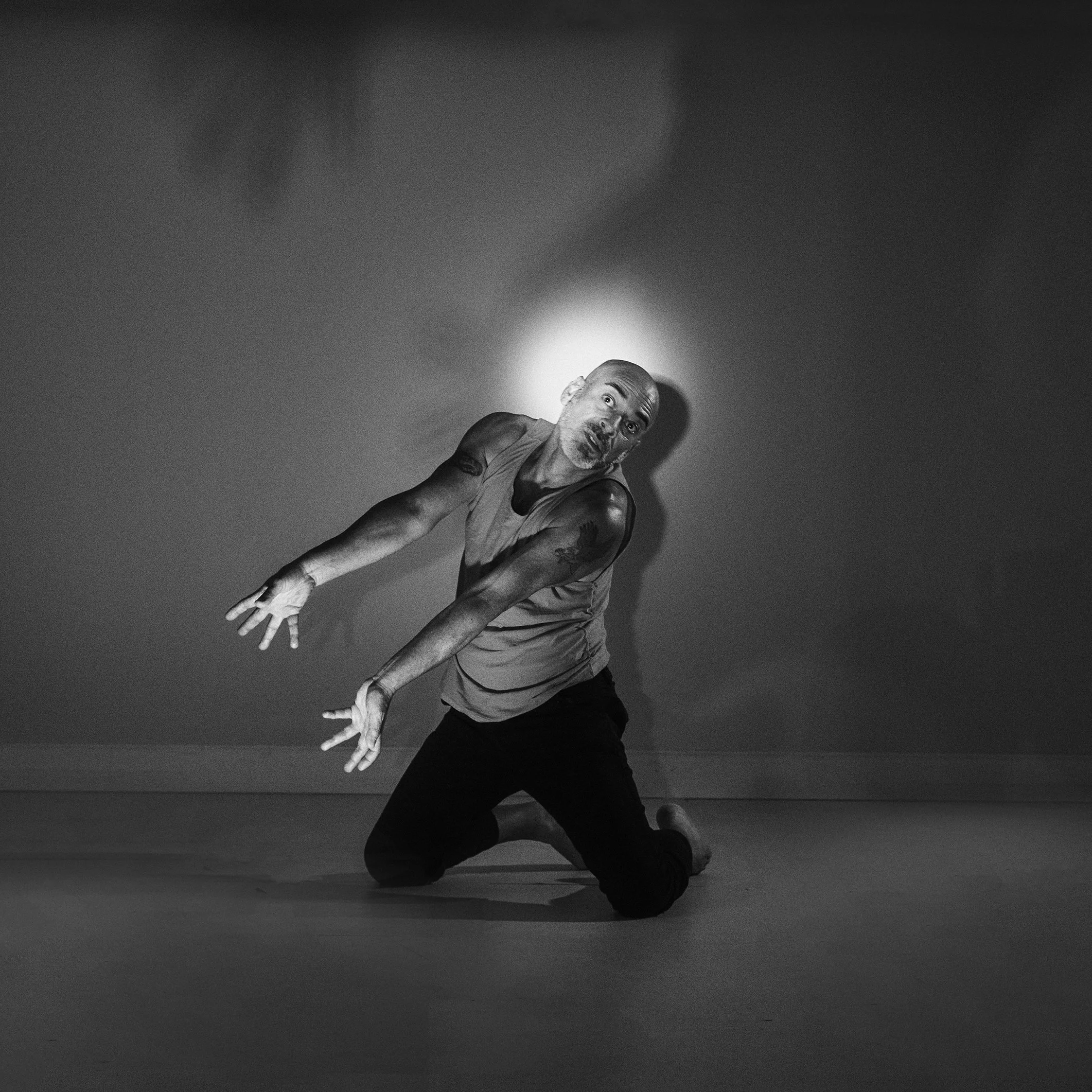 Homem segurando-se no chão, expressão facial de surpresa ou emoção, em cenário com fundo escuro e iluminação focada.