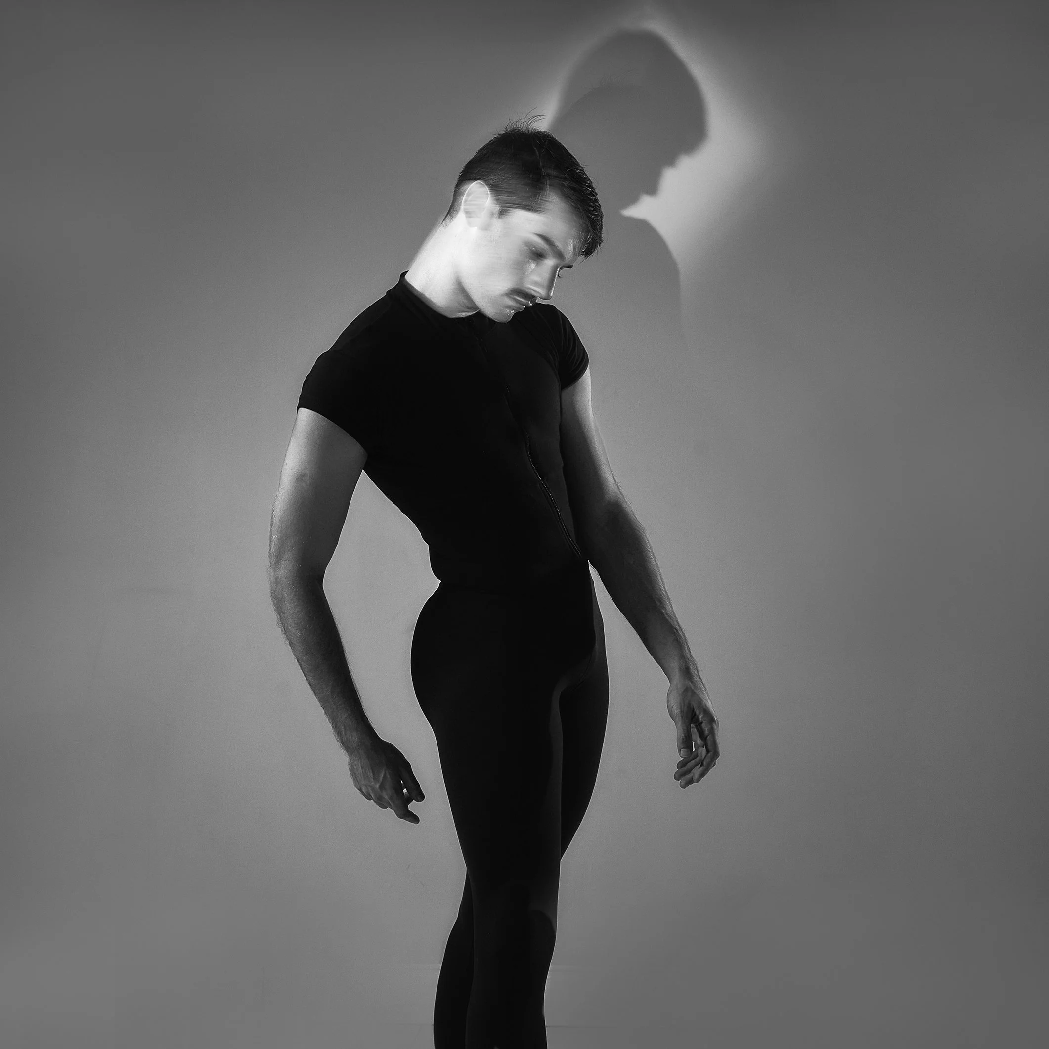 Homem em roupas pretas, olhando para baixo, com sombra projetada na parede atrás dele.