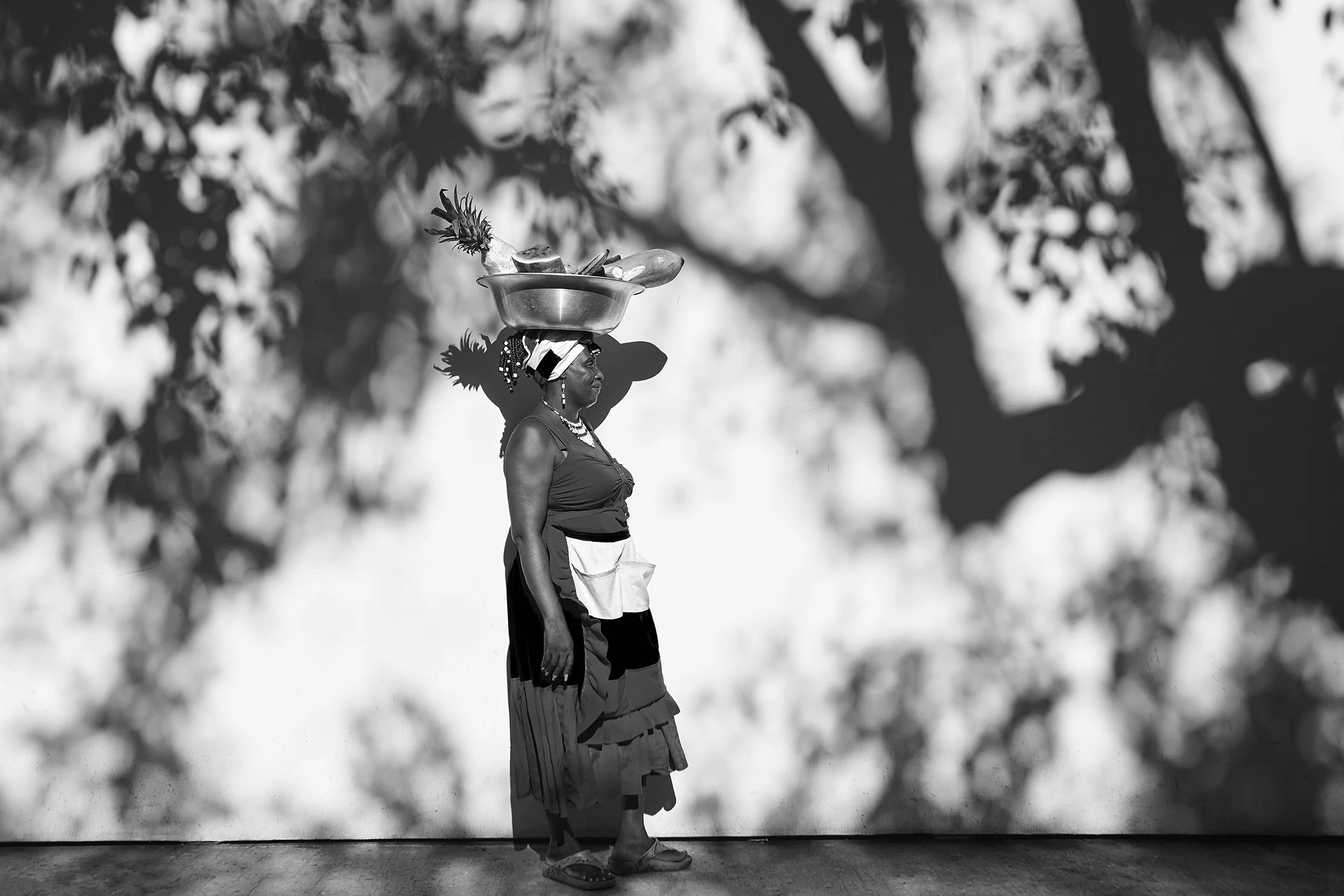 Mulher indígena caminhando com uma cabeça carregando uma vasilha com frutas e plantas, usando trajes tradicionais e acessórios de cabeça, ao lado de uma parede com sombra de árvores.