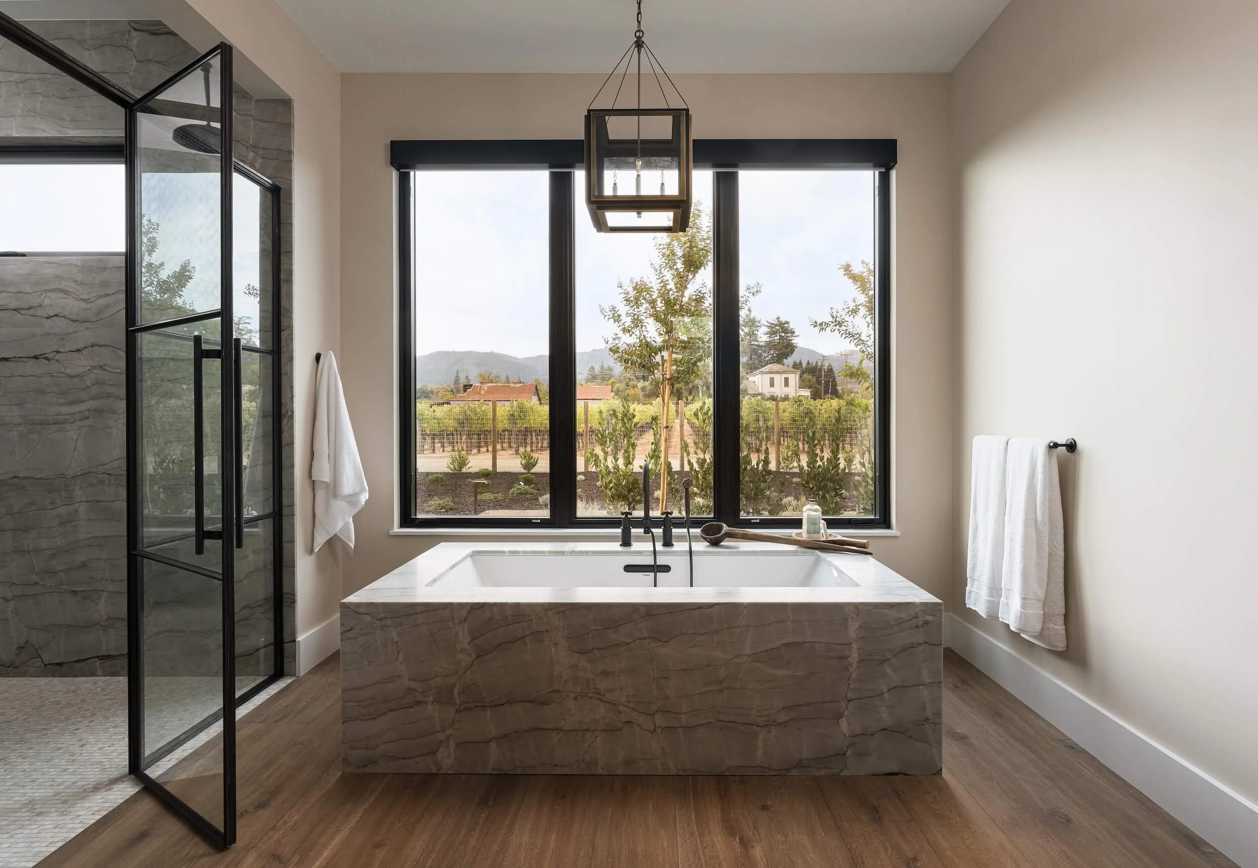 Elegant master bathroom lighting featuring a modern chandelier over a soaking tub with automated window shades.