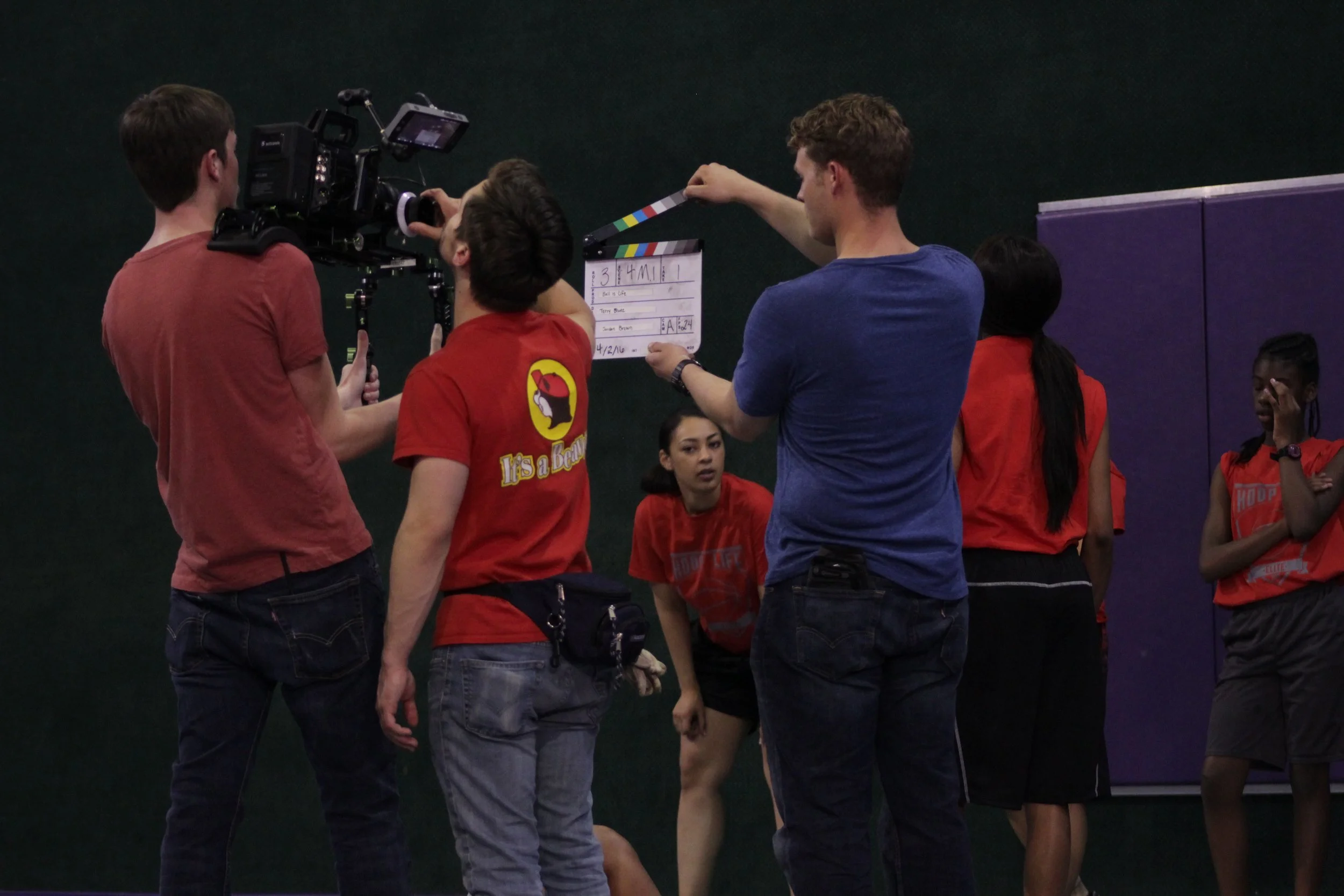 A group of people on a film set, including a cameraman, a clapperboard operator, and several others, at an indoor basketball court.