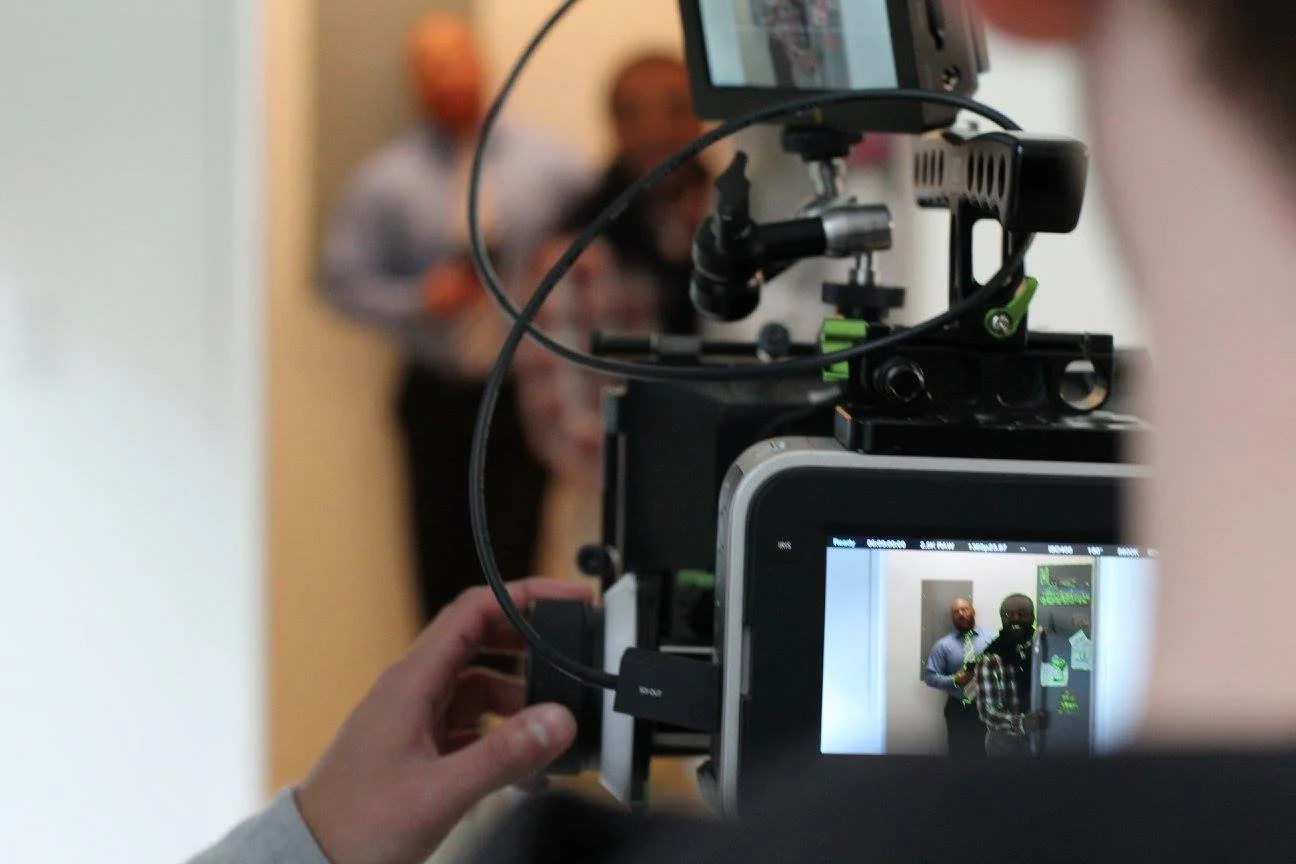 A camera on a tripod filming two men in a room with beige walls and orange vases in the background.