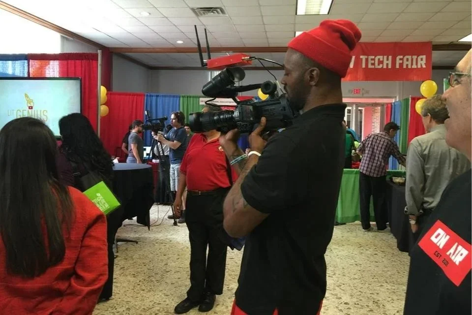 A person with a red beanie holding a professional video camera at a tech fair with several booths and attendees in the background.