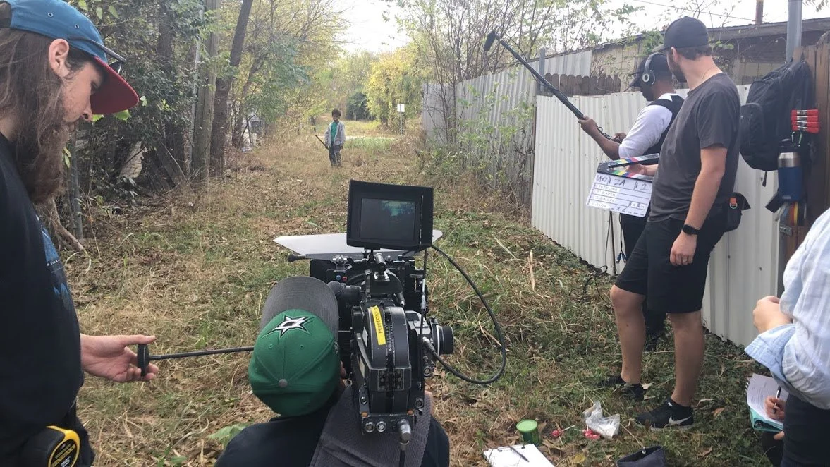 Filming crew on location filming a scene in a grassy outdoor area with trees and a fence, with a young person in the background.
