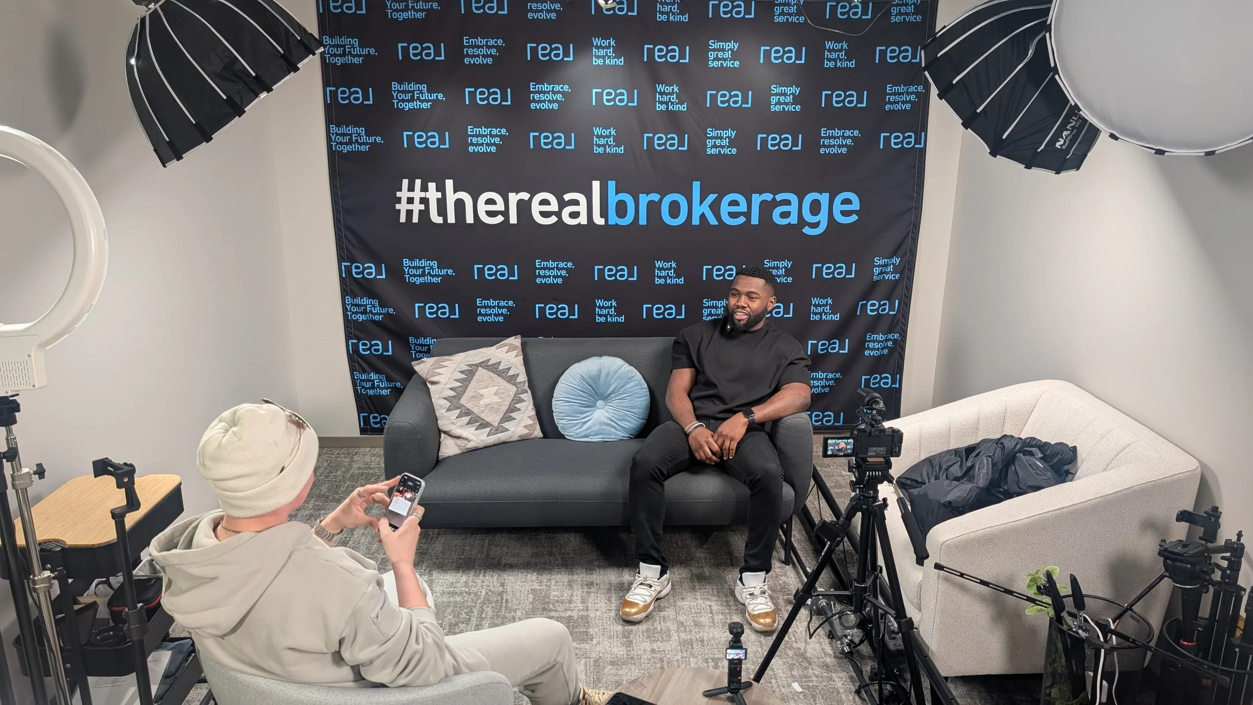 An interview setup with two men, one on a gray sofa and the other on a white chair, in front of a black backdrop with blue and white text and hashtags related to brokerage. There is professional filming equipment and lighting around them.