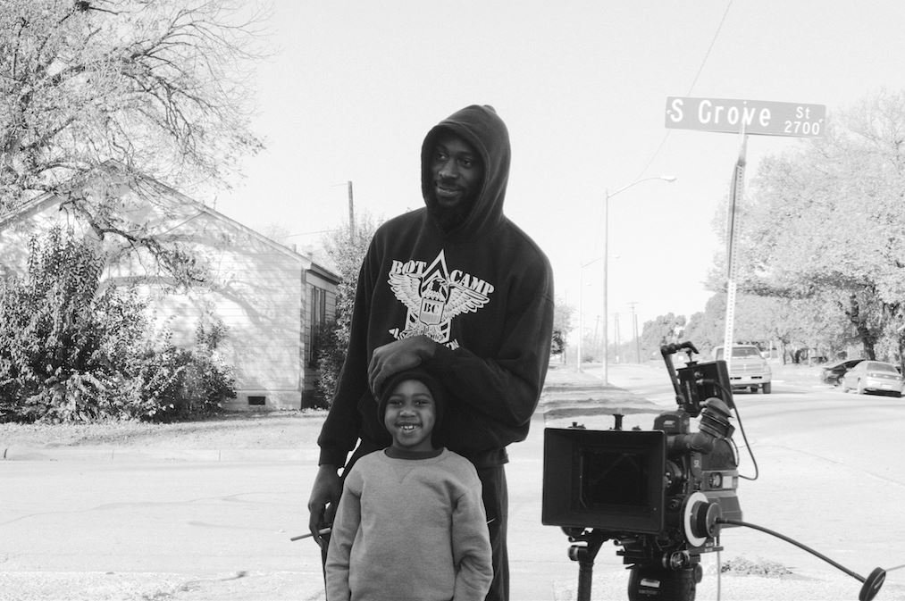 A man and a young girl standing outside near a camera on a tripod. The man is wearing a hooded sweatshirt and the girl is smiling. There is a street sign that reads "S Grove St" in the background.