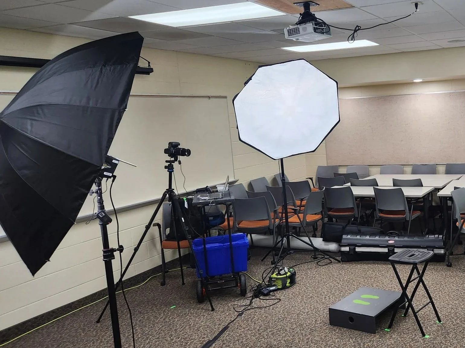Photography or video studio setup with lighting equipment, a camera on a tripod, and tables with chairs arranged in a room with beige walls and ceiling.