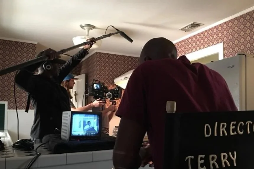 Filming crew capturing a scene in a kitchen for a video or film production, with a cameraman, a person holding a boom microphone, and a man with a shaved head working at a counter.