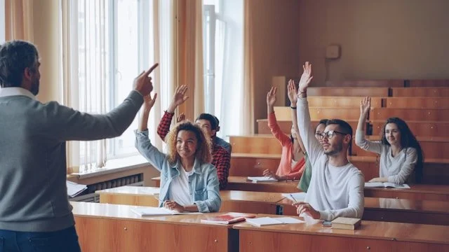 Teacher conducting a classroom quiz with students raising their hands.