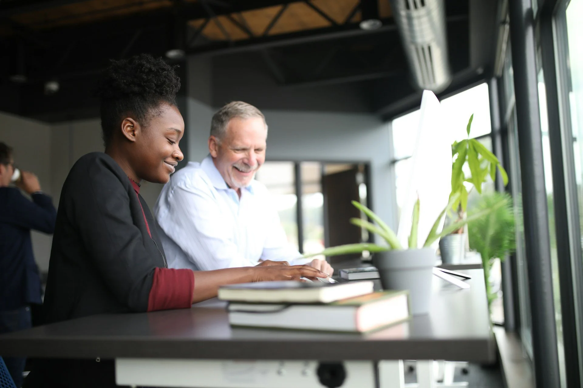 Two people sitting at a table near a window, smiling and working together with books and a potted plant on the table.