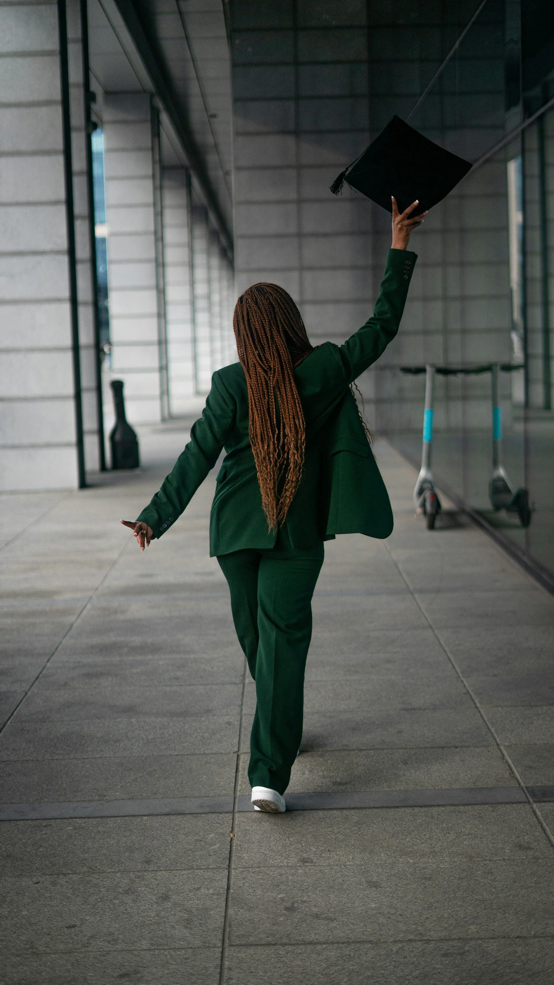 A person with braided hair, wearing a green suit and white shoes, walking outside on a modern urban sidewalk, raising a graduation cap in the air.