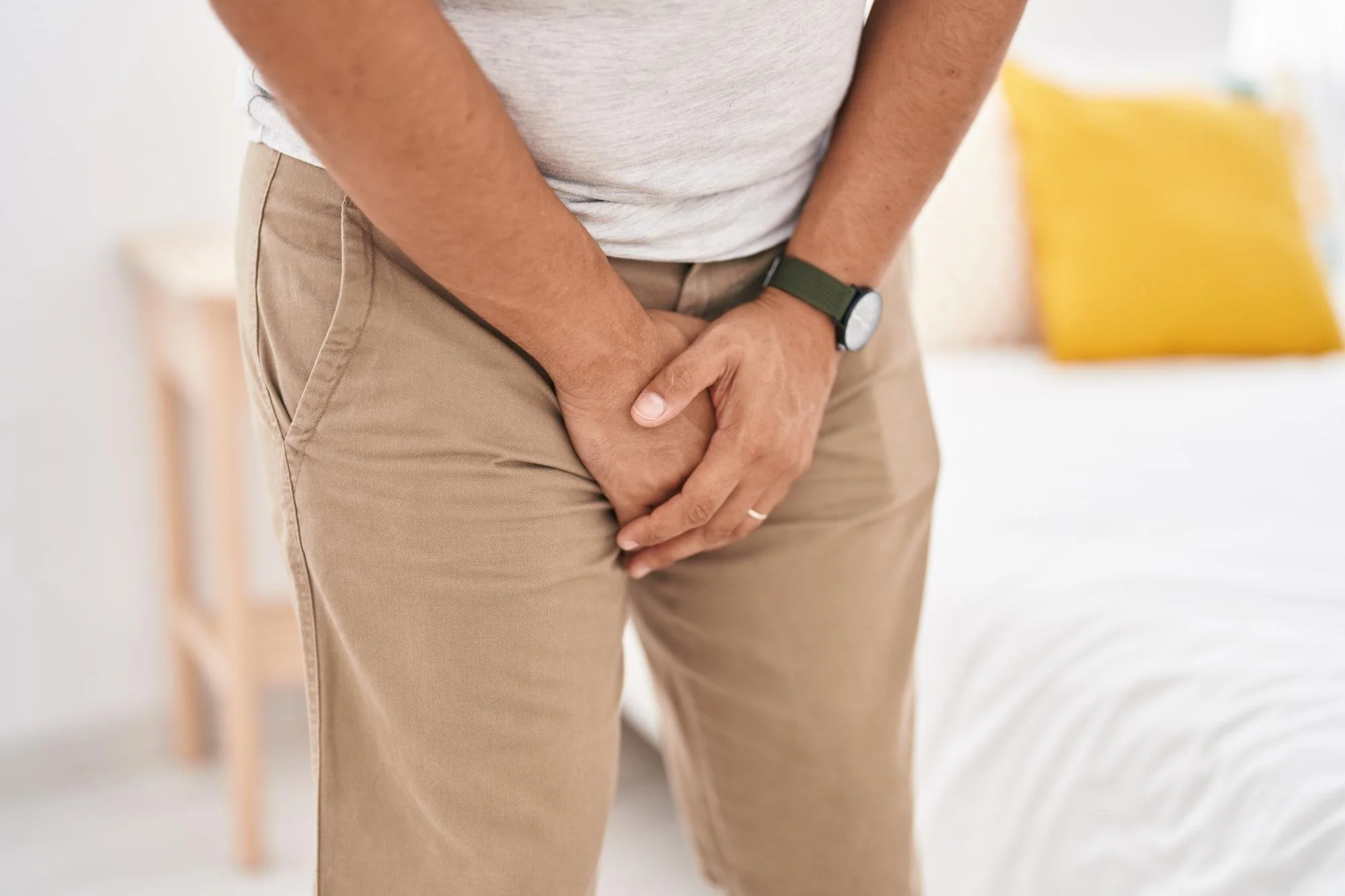 A man in beige pants and a light gray shirt with a watch on his wrist, holding his lower abdomen area with both hands.