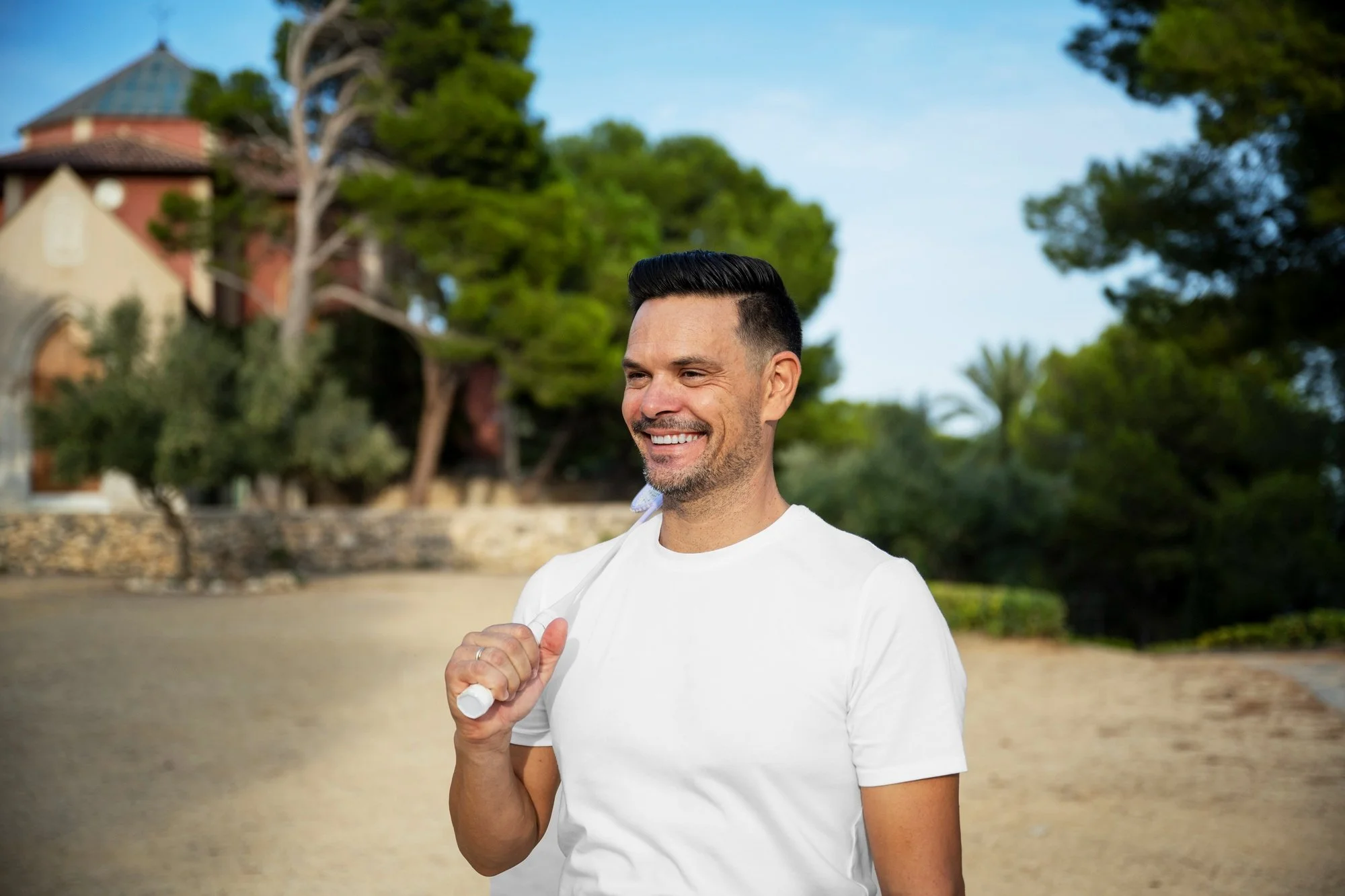Smile of a young man holding a rolled-up paper in a white t-shirt with trees and buildings in the background.